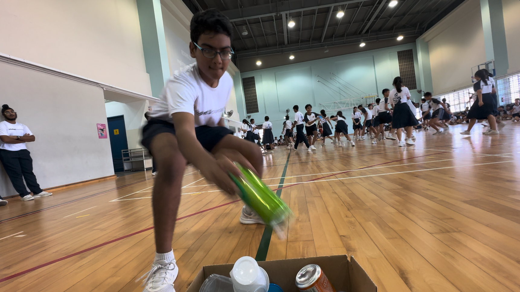 A boy with glasses tosses a green bottle into a cardboard box on a gymnasium floor during a school activity.