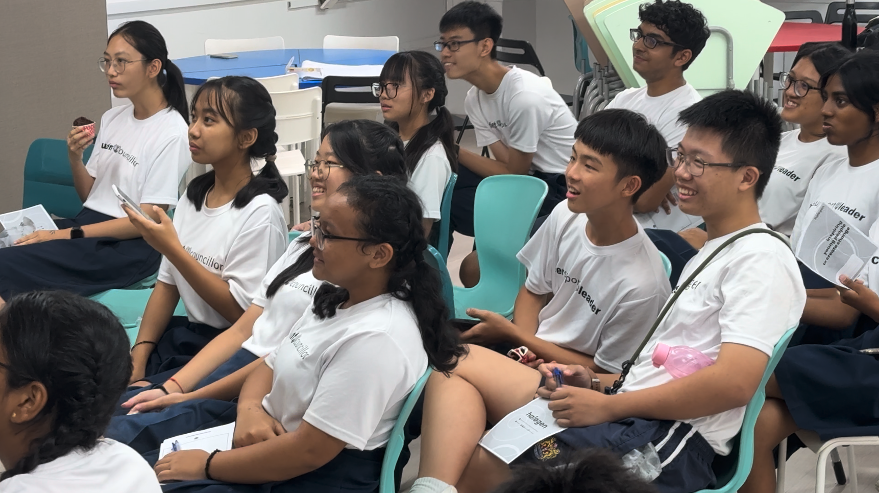 A group of teenagers in white matching t-shirts sit on teal chairs in a classroom, smiling and holding papers.