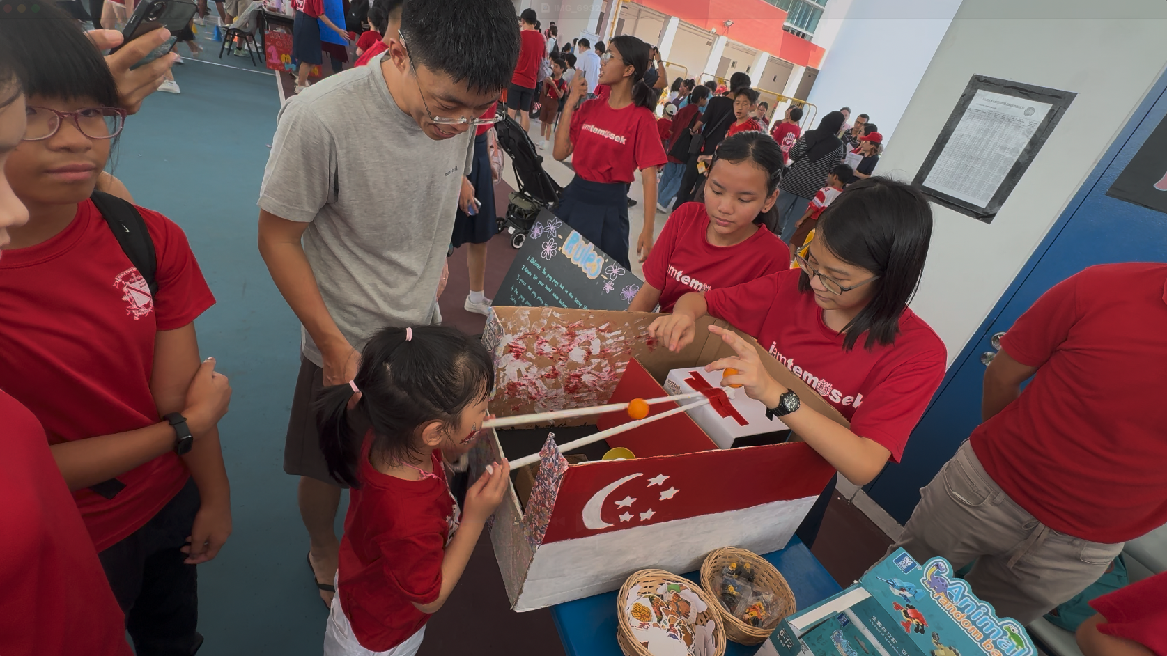 Children and adults in red shirts play a chopstick-and-ball game at a school event booth decorated with a Singapore flag.