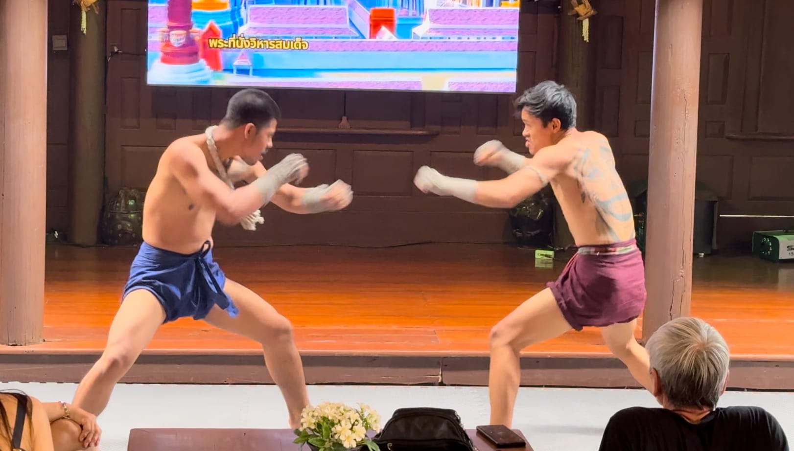 Two shirtless men perform a traditional Muay Thai demonstration on a wooden stage before an audience.