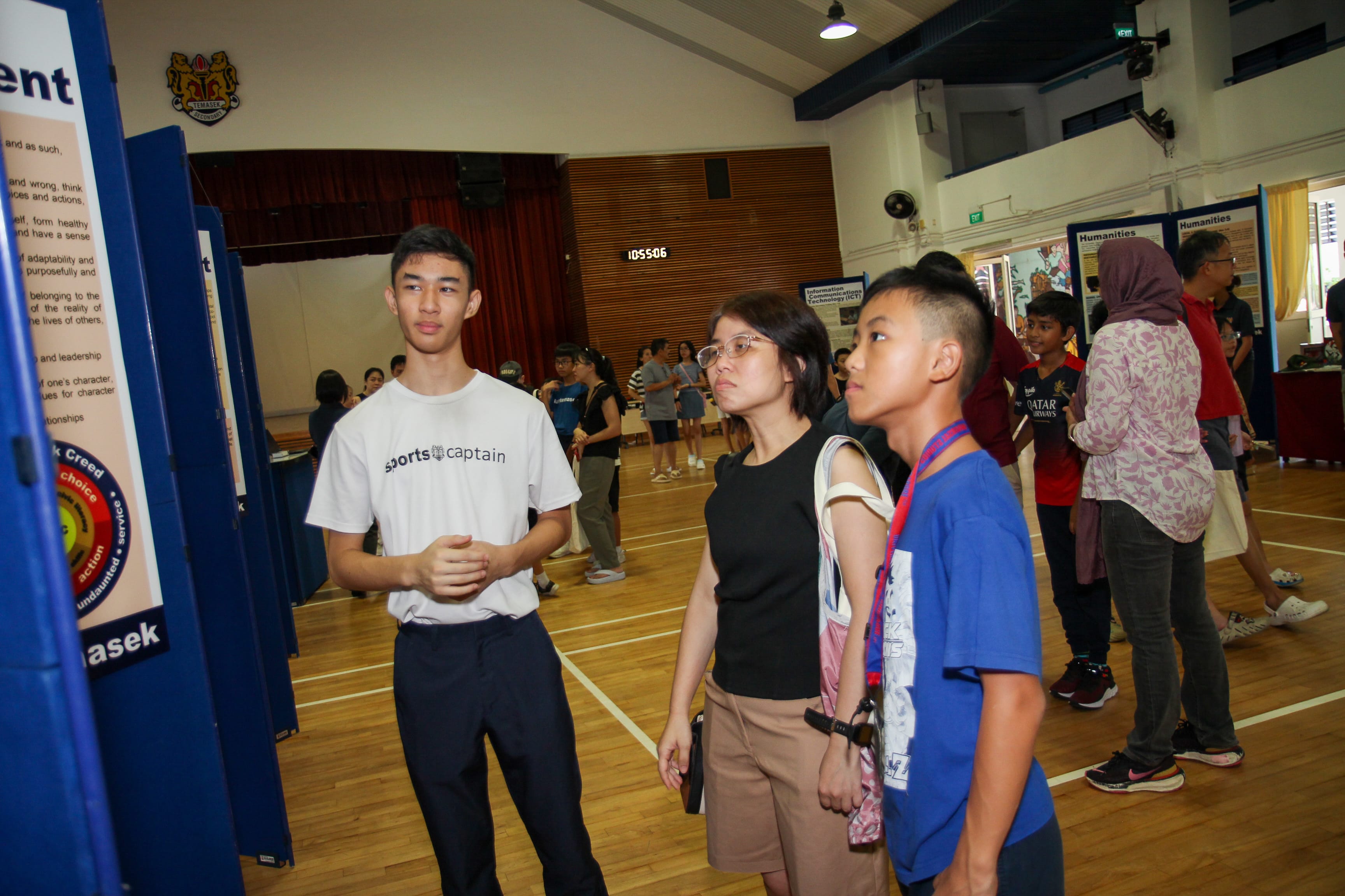Three people stand in a gym, looking at display boards, with other visitors in the background.
