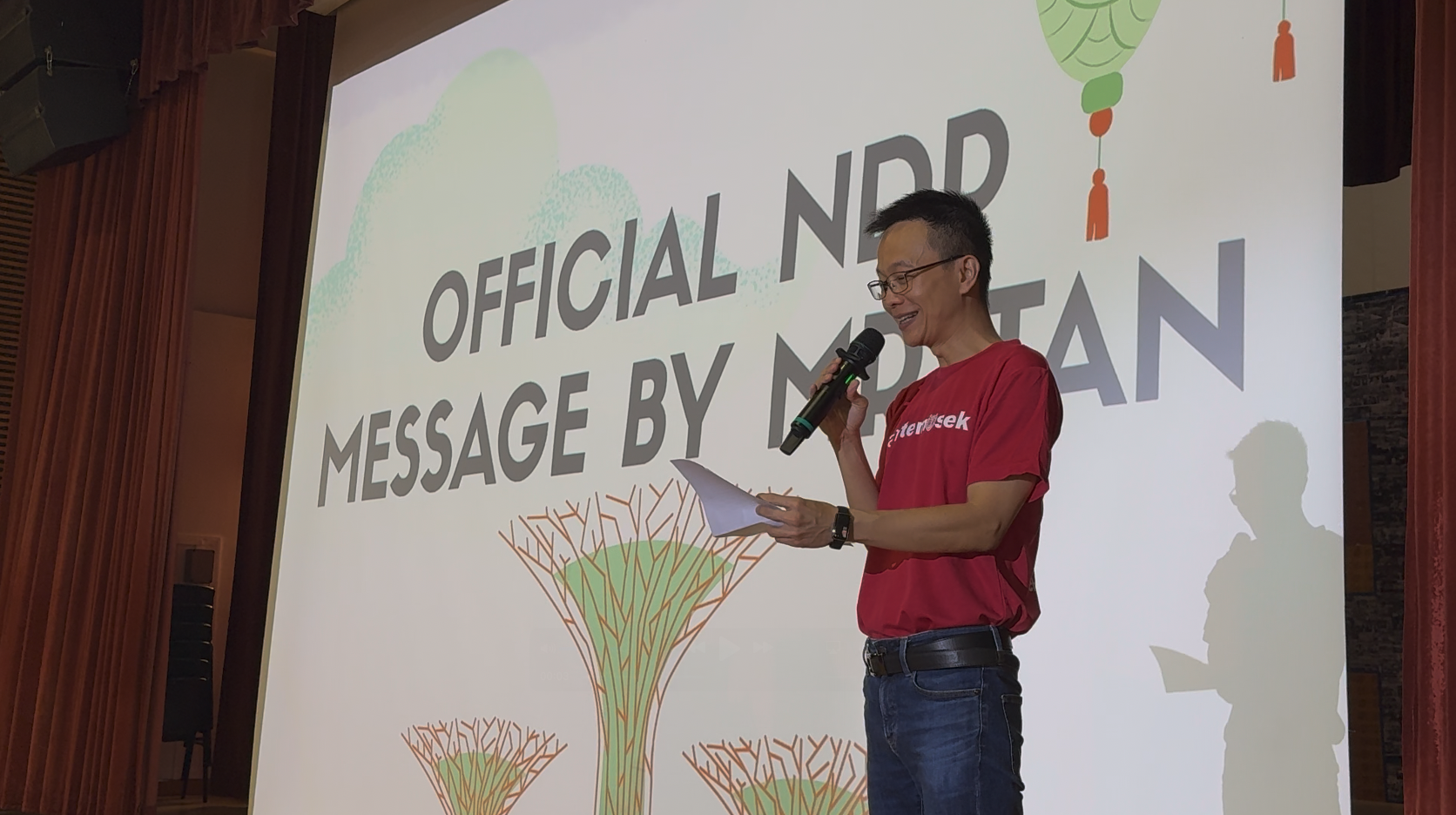 Man in red shirt holding microphone and papers speaks in front of a projection reading "Official NDP Message by Mr Tan."