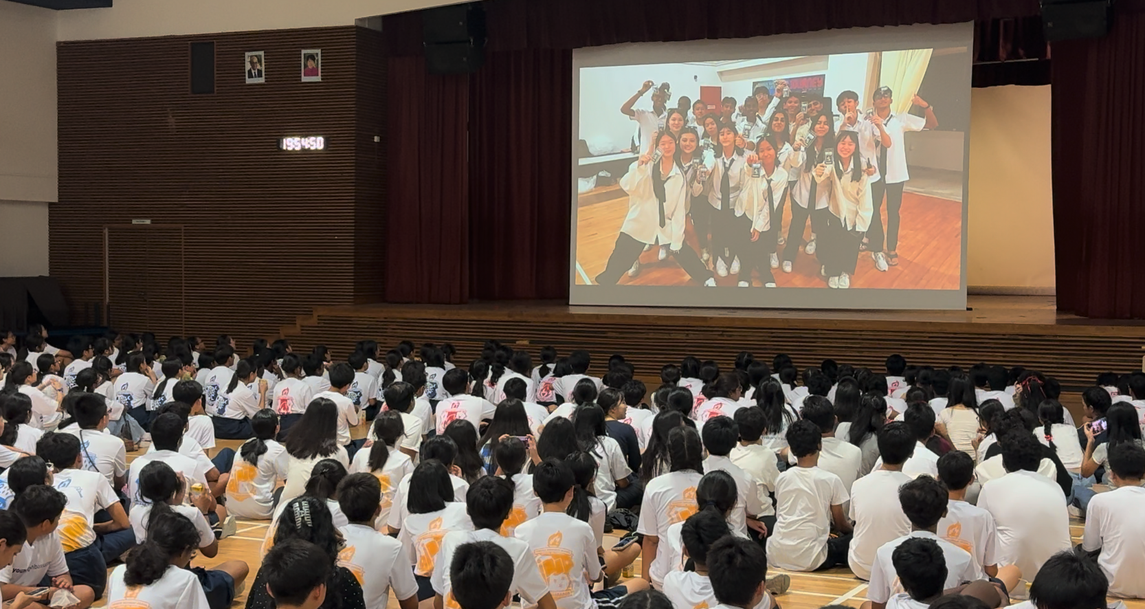 Large group of students in white t-shirts sitting on a gym floor, watching a projected group photo on a stage screen.