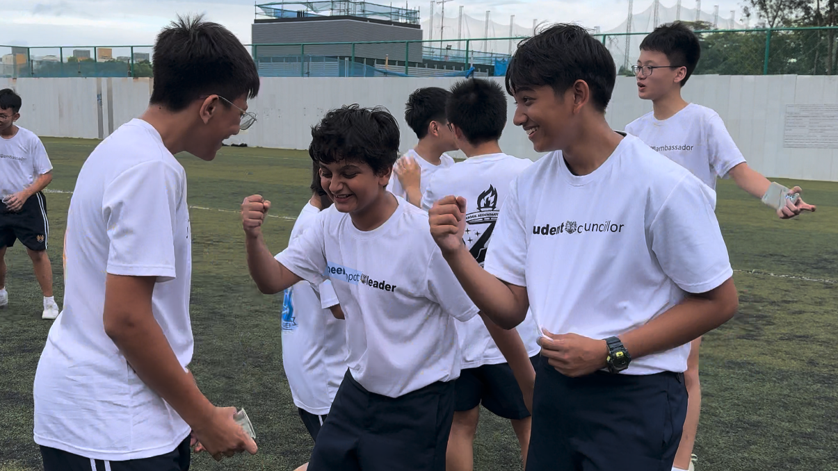 Students in white school role T-shirts celebrate on an outdoor sports field, smiling and raising fists.