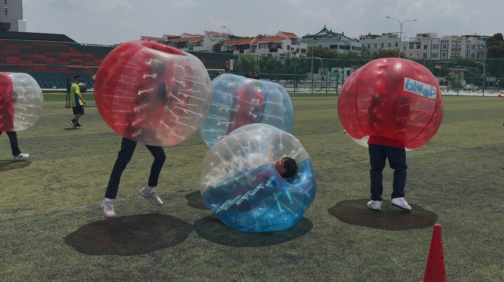 People wearing large inflatable bubble balls in red and blue play on a grassy field, with one person fallen over.