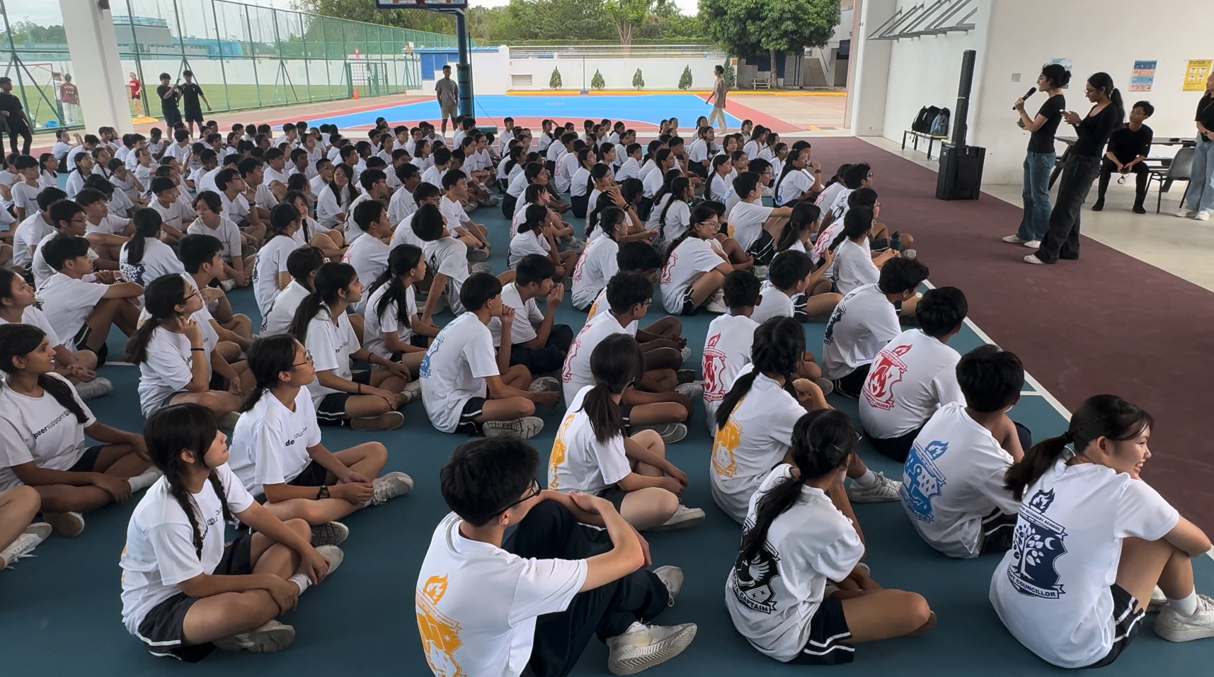 Large group of students in white shirts sitting on a court floor, facing presenters speaking with microphones.