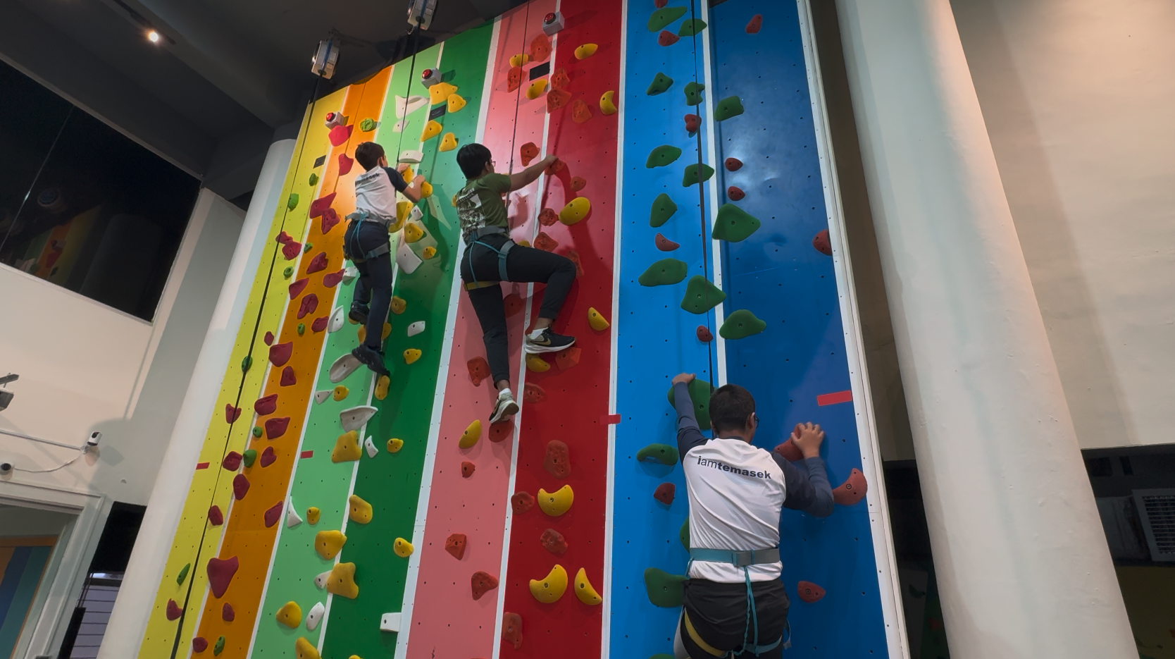 Three people wearing harnesses climb a tall indoor rock wall with colorful striped panels and varied holds.