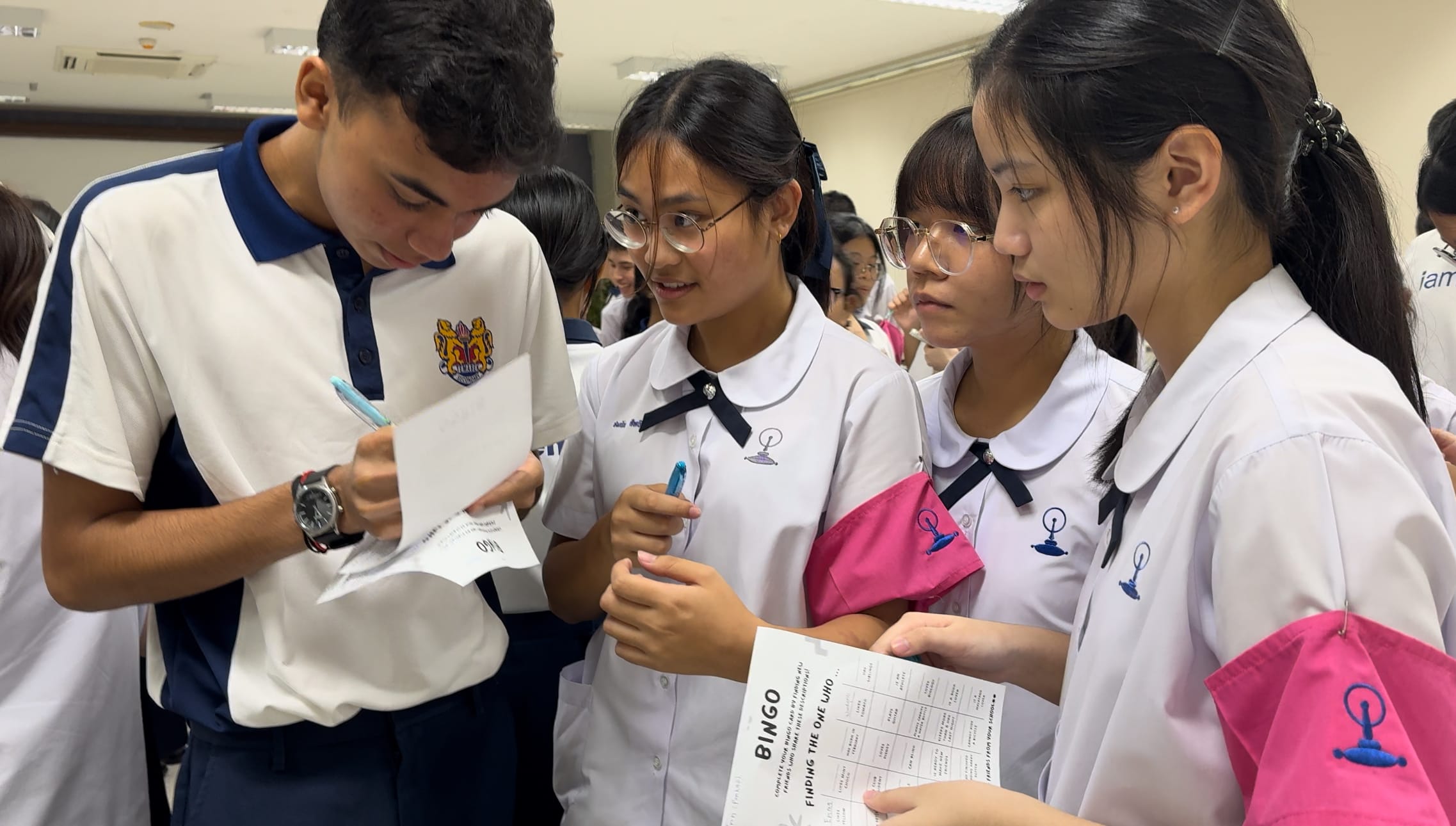 Four students in school uniforms huddle together, writing on papers including a bingo sheet titled "Finding the One Who."
