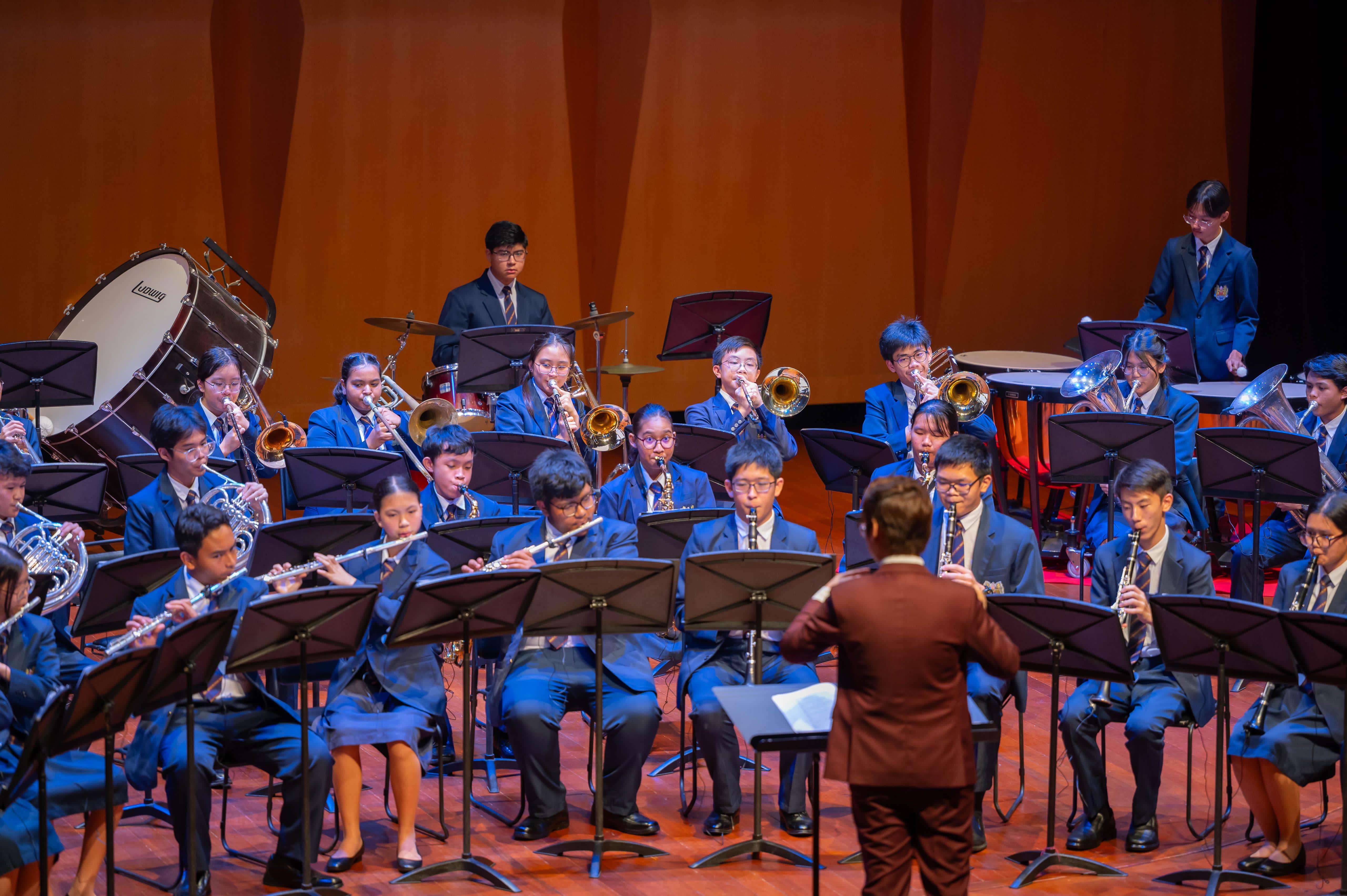 A conductor in a red suit leads a youth wind band in blue uniforms performing on a warm-lit stage.