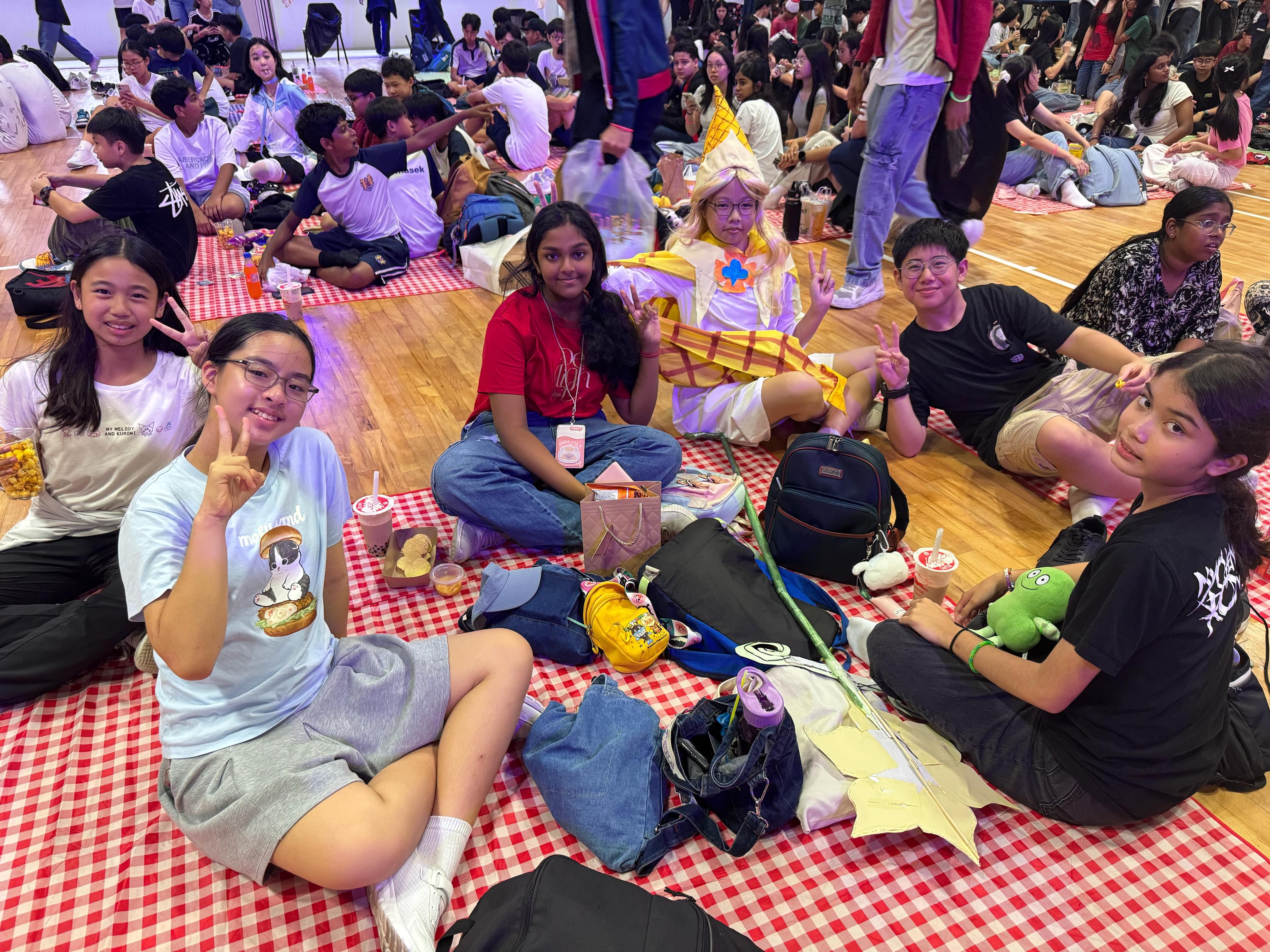 A group of teens sit on red checkered blankets on a gym floor, smiling, one wearing a costume with a tall hat.