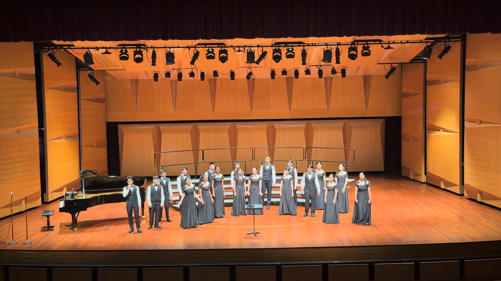 A choir in gray formal attire performs on a lit wooden stage with a grand piano and overhead stage lights.