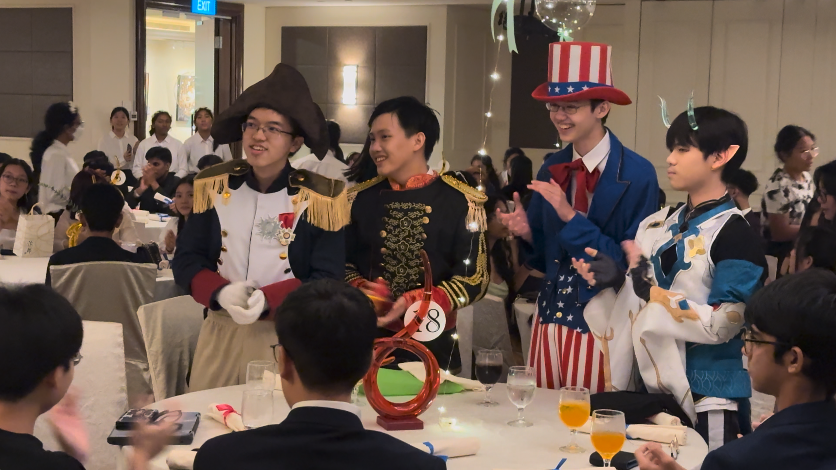Four young men in costumes including Napoleon and Uncle Sam stand smiling at a banquet hall event.