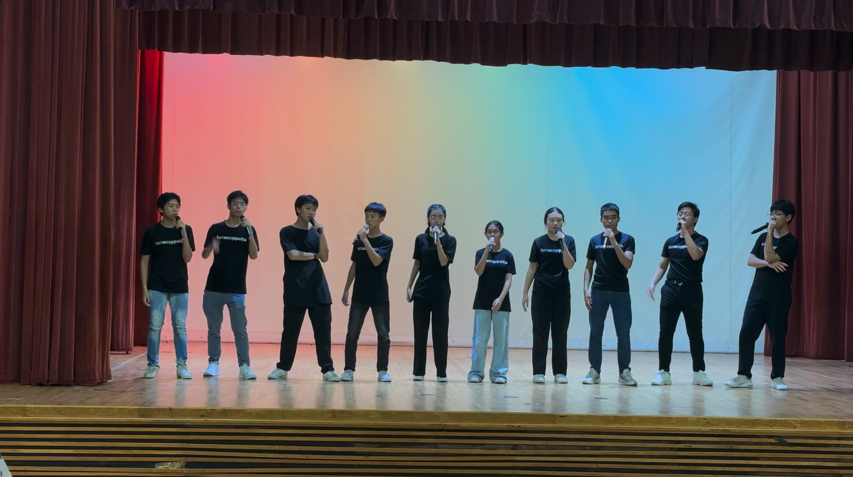 Ten young performers in black t-shirts stand in a row on stage holding microphones before a colorful backdrop.