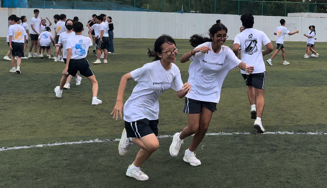 Two girls in white t-shirts laugh and run together on a grass field during a school sports event.