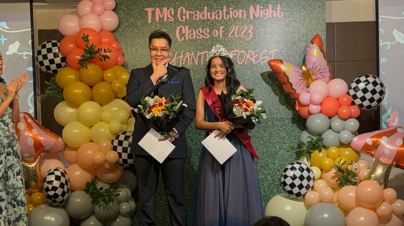 Two students hold bouquets at TMS Graduation Night Class of 2023, surrounded by colorful balloons; a girl wears a crown and sash.