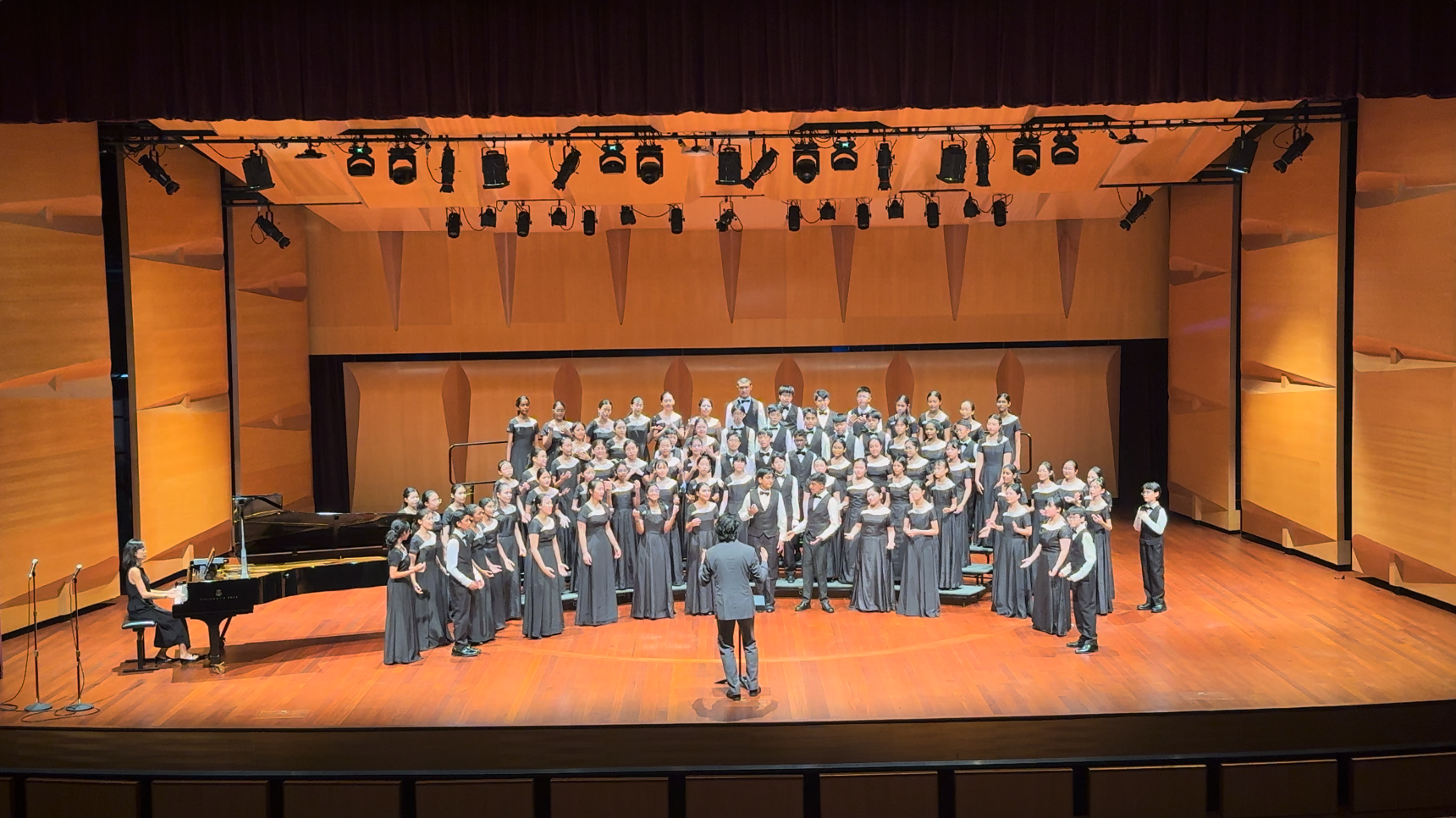 A large youth choir in dark uniforms performs on a lit concert stage with a conductor and pianist.