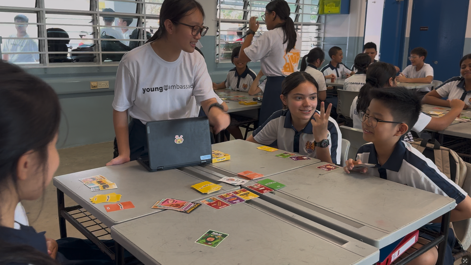 Students in school uniforms play a card game at a table while a young ambassador in a white shirt facilitates.