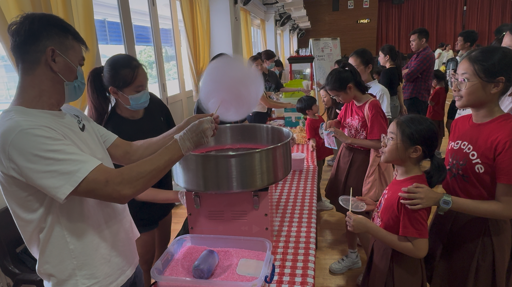 Two masked adults make cotton candy at a machine while children in red shirts watch and wait in line indoors.