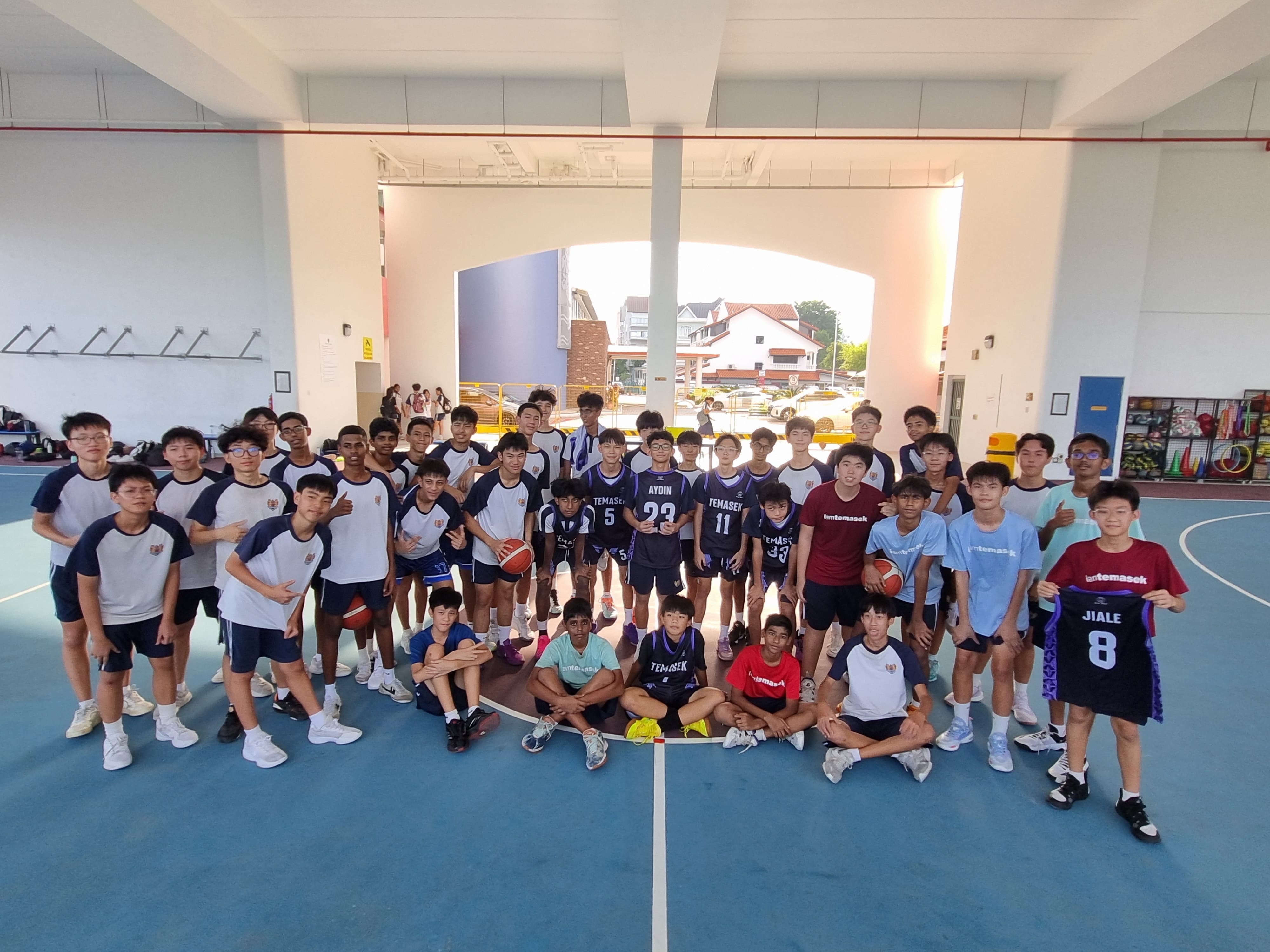 A large group of teenage boys in basketball uniforms pose together on an indoor court at Temasek school.