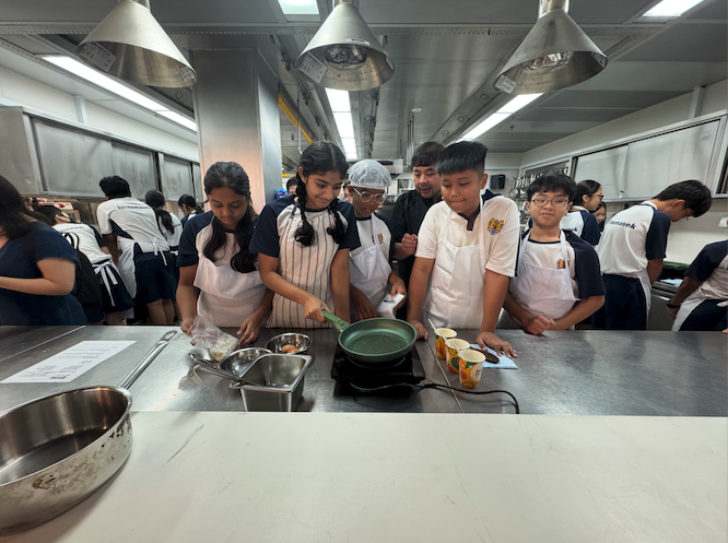 Students in aprons gather around a stainless steel counter, watching a classmate cook in a green pan on an induction stove.