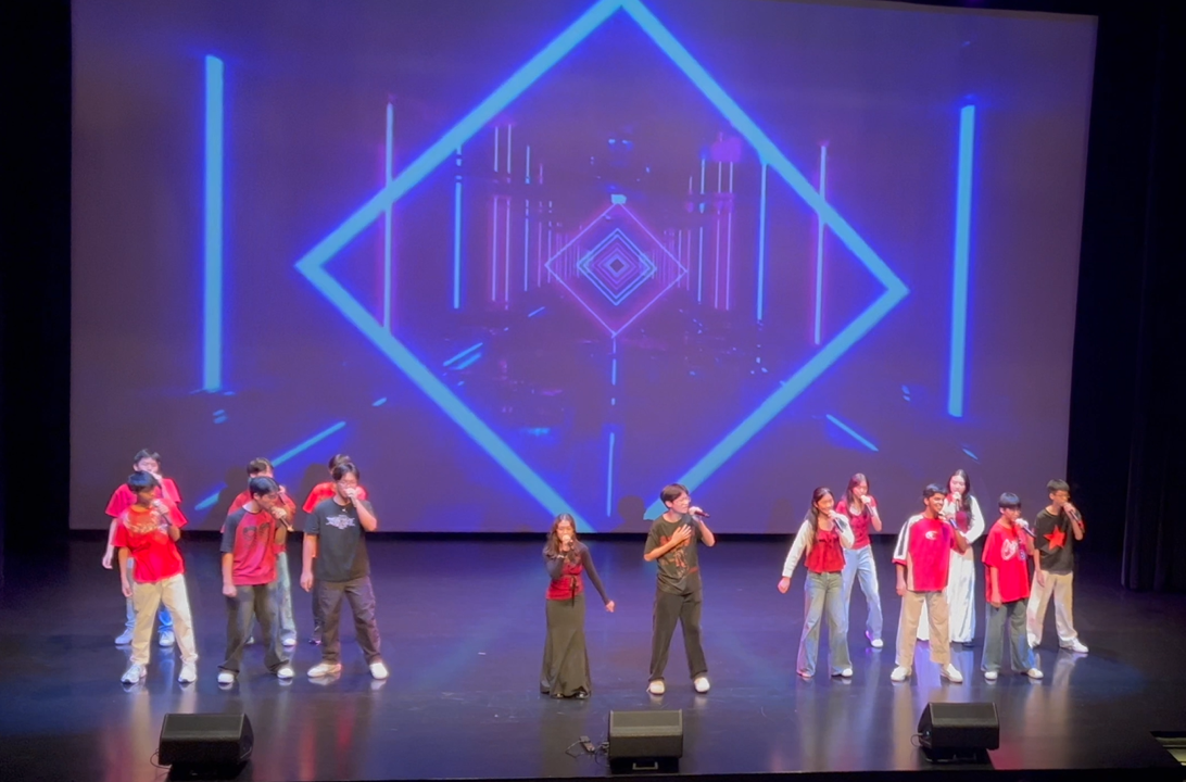 A group of young performers in red and black outfits singing on stage with a blue geometric projected backdrop.