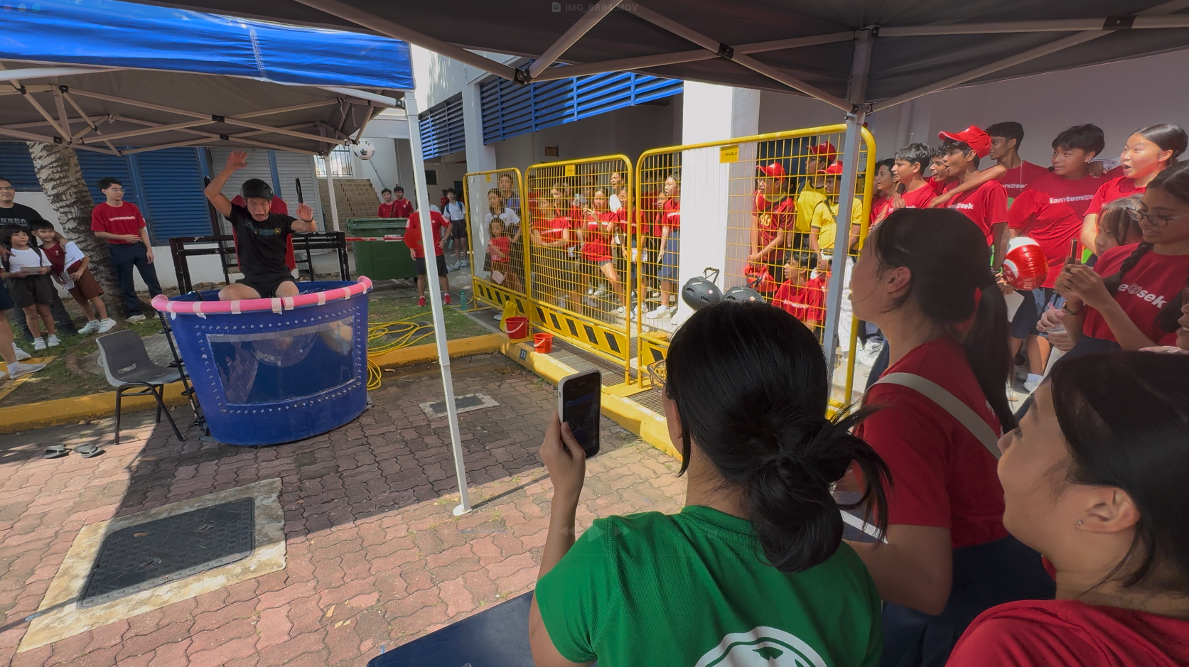 A crowd watches a dunk tank activity outdoors, with a person sitting above a blue water tank at an event.