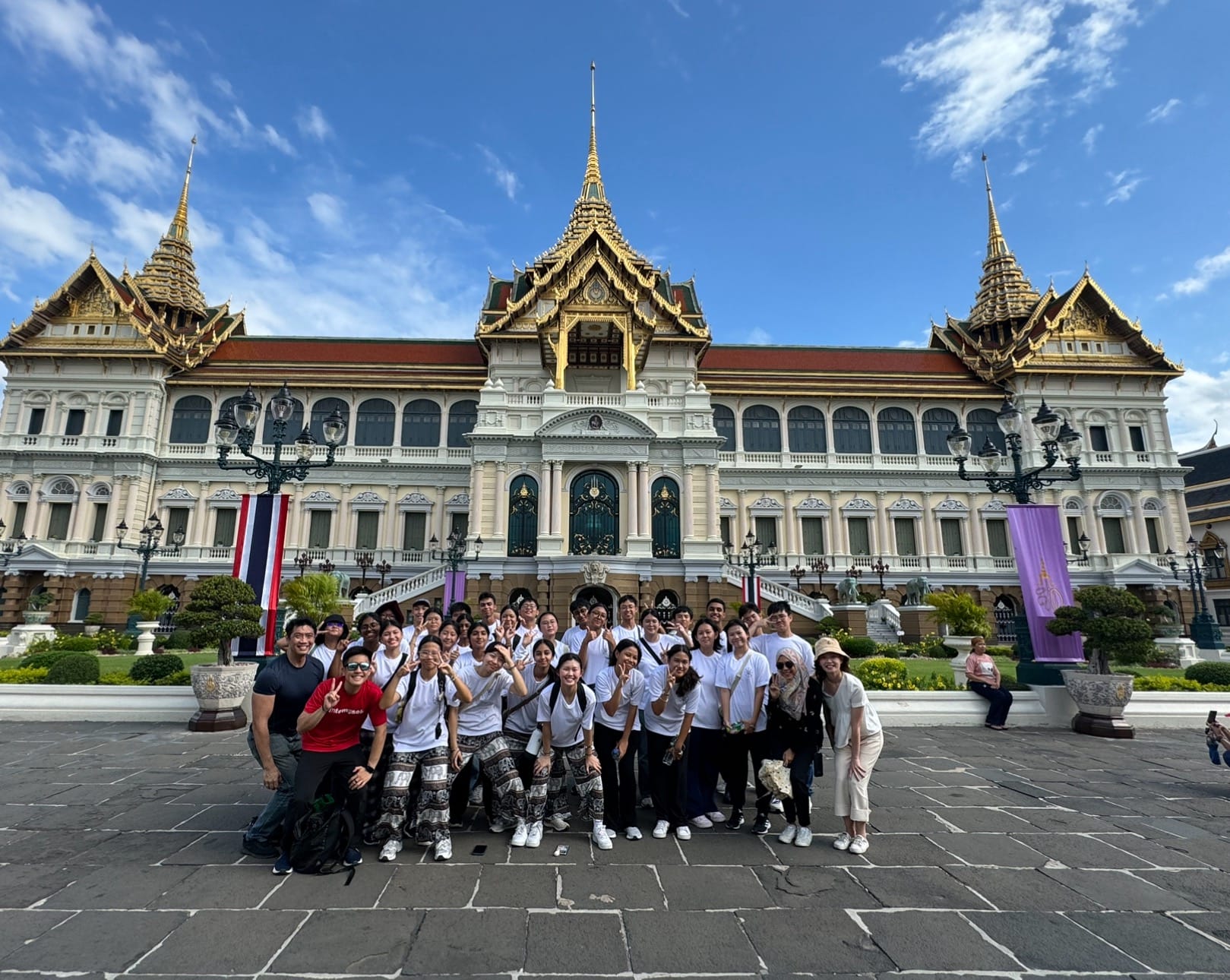 A large group of tourists poses in front of the Grand Palace in Bangkok, Thailand, under a blue sky.