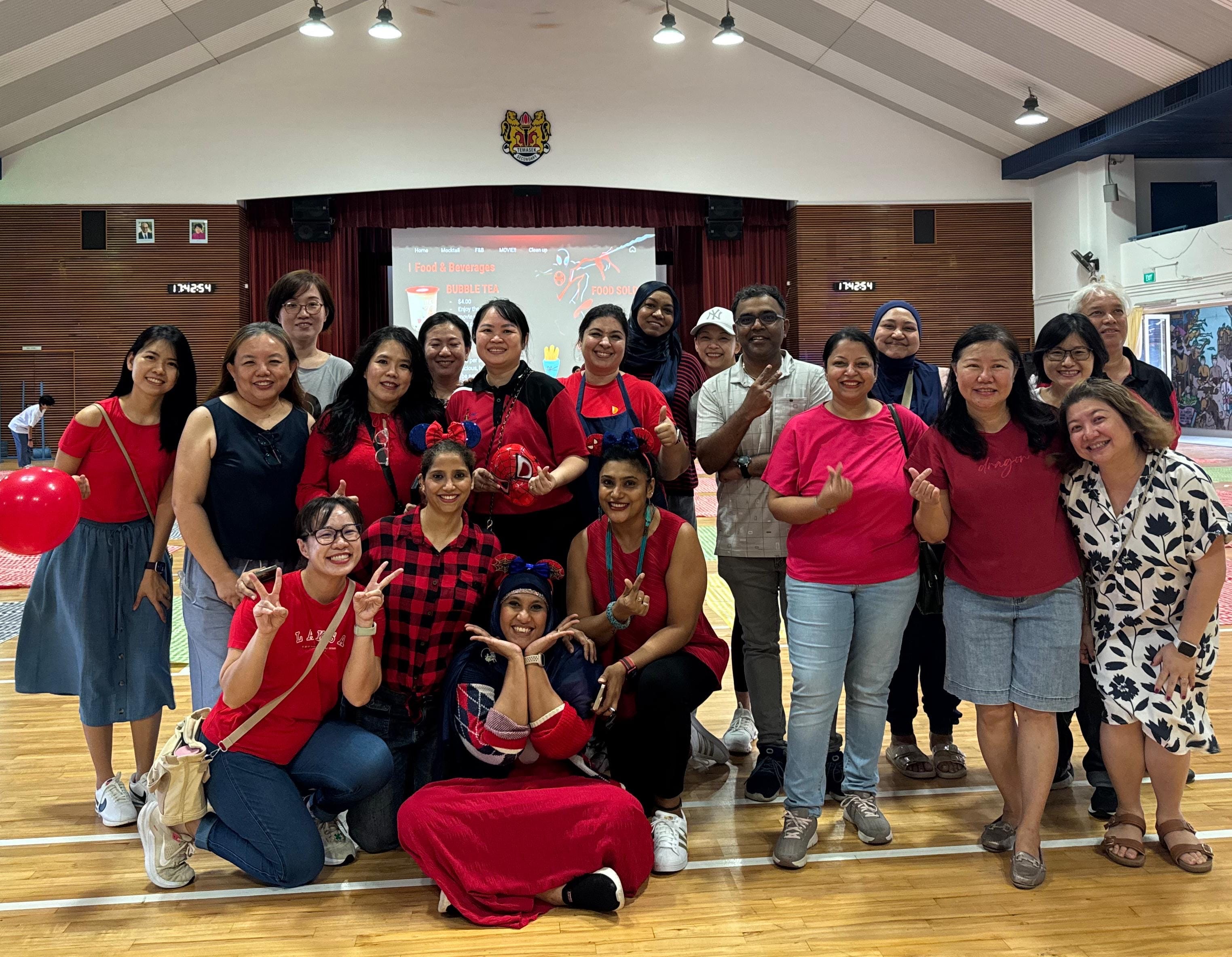A large group of smiling people mostly wearing red pose together in an indoor hall with a projection screen behind them.