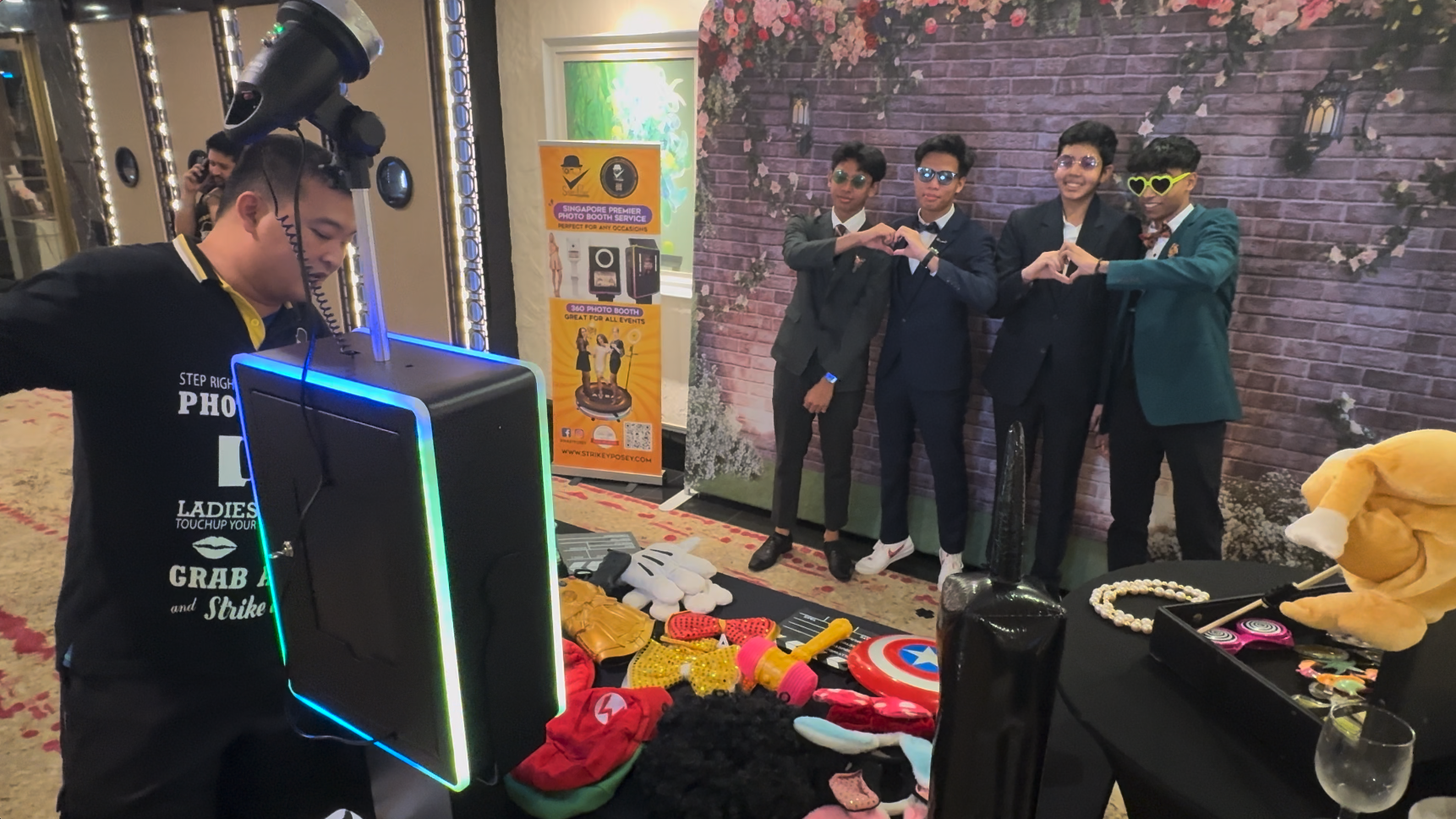 Four young men in suits pose making heart shapes at a photo booth with props and a floral backdrop.