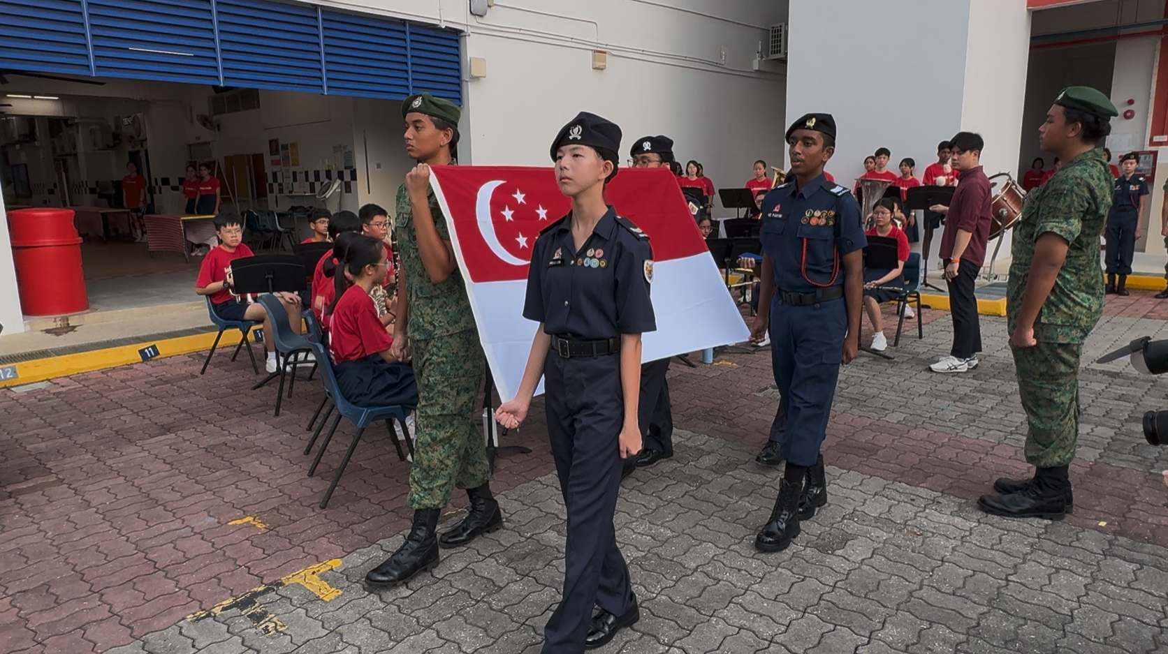 Students in uniform carry the Singapore flag during a school ceremony, with a band seated in the background.