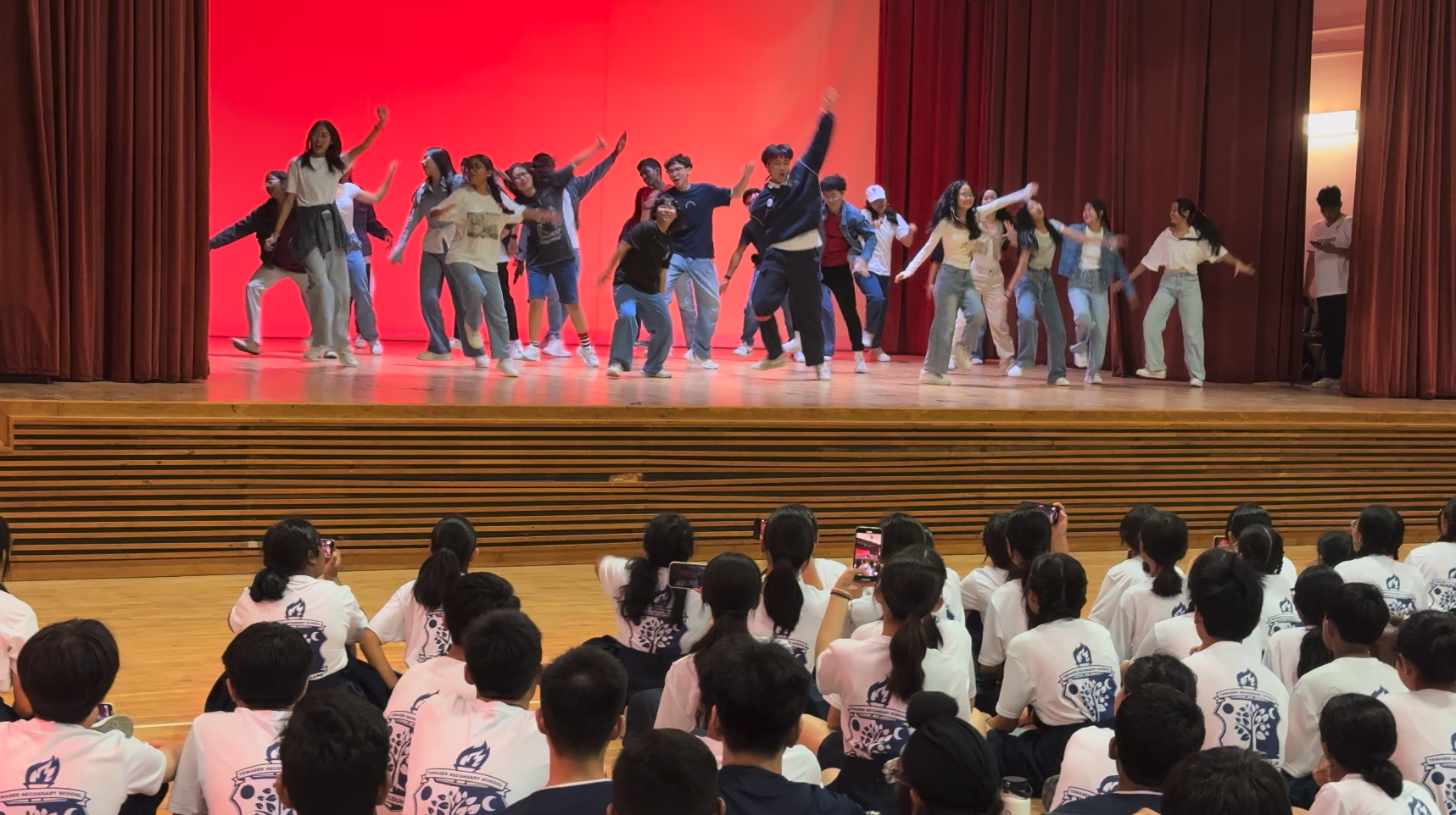 A large group of students dancing on a red-curtained stage while uniformed students watch from the floor.