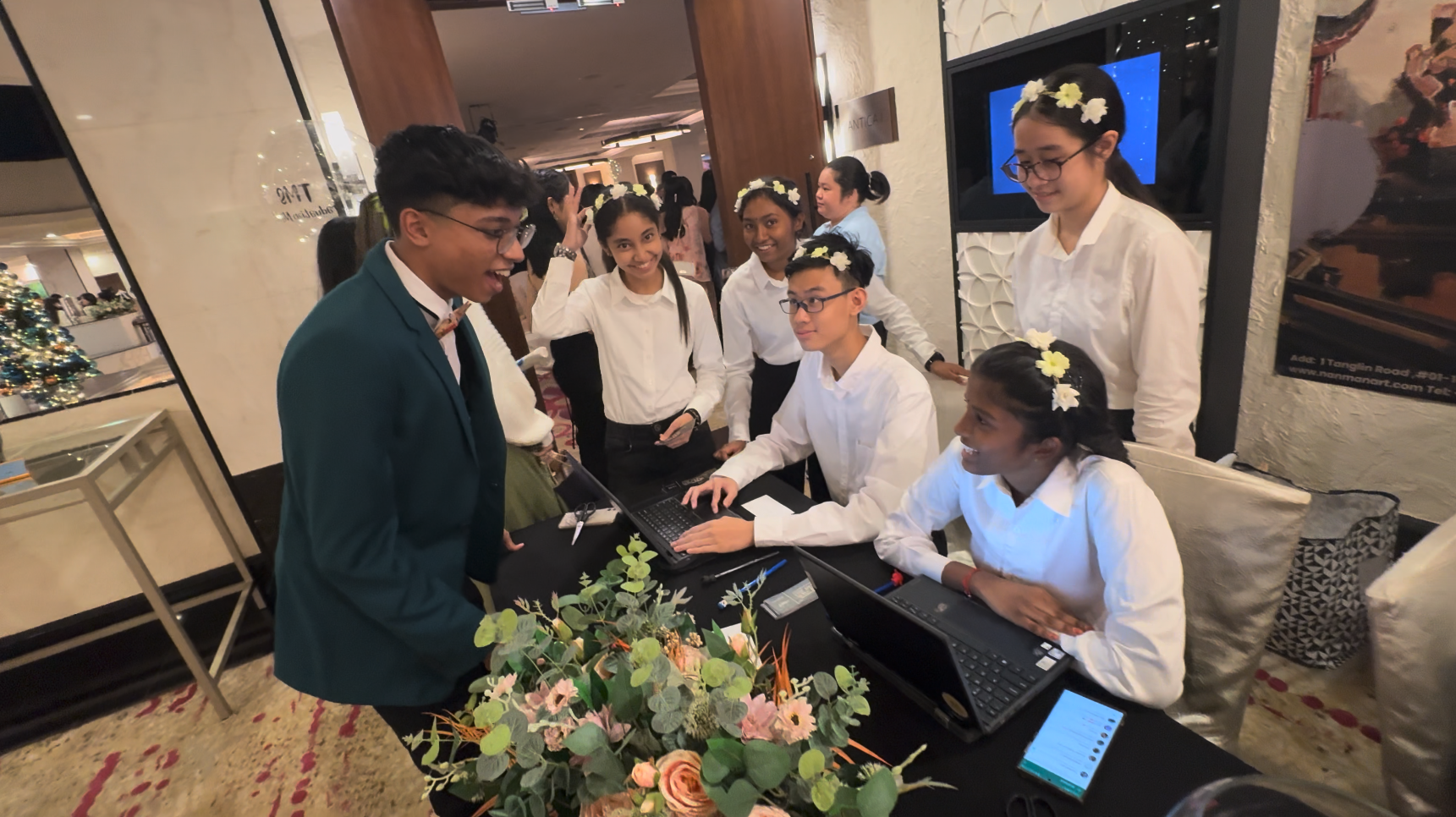 A young man in a teal blazer talks with several teens in white shirts at a registration desk with laptops and flowers.