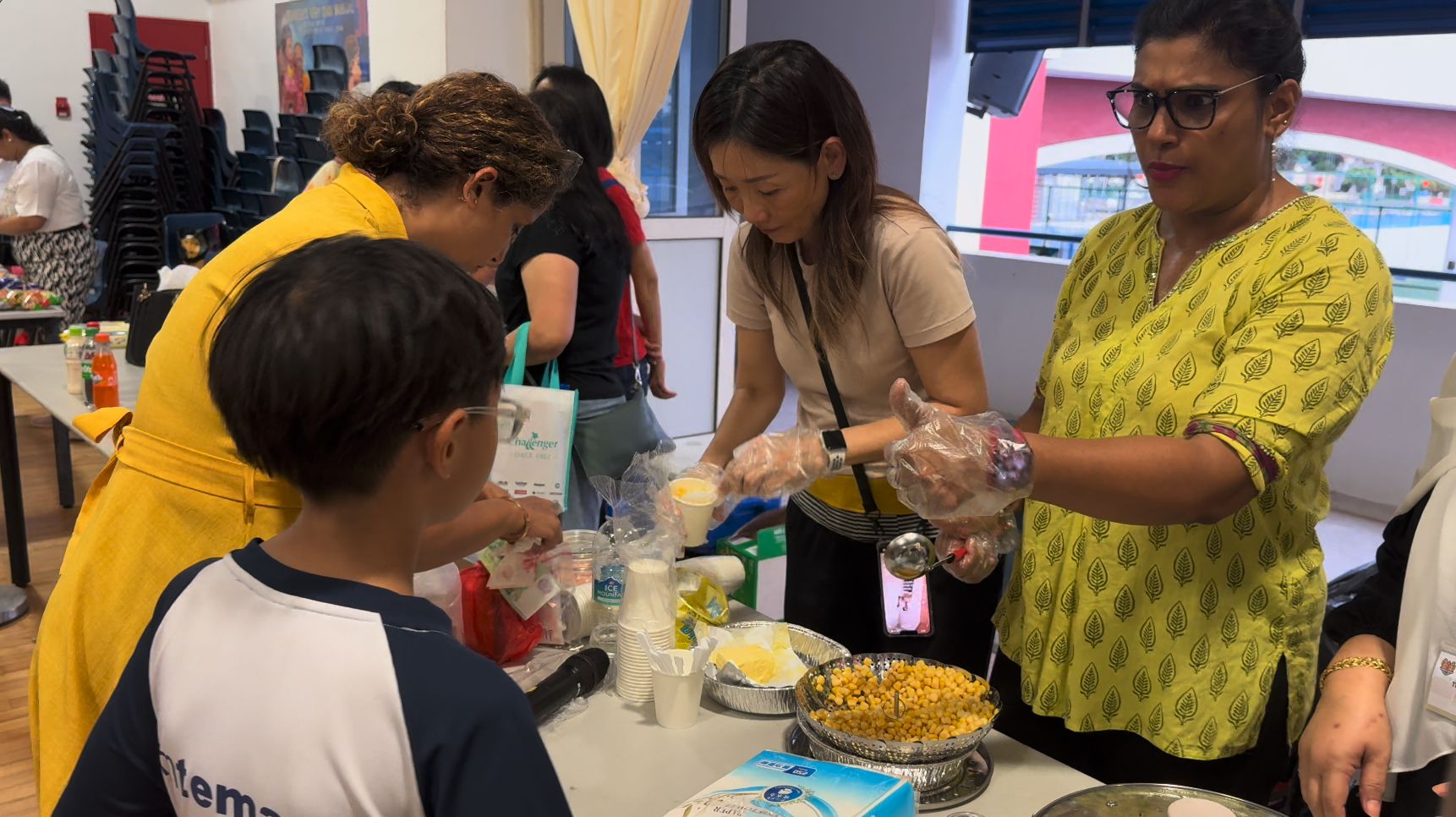Several people gather around a table serving food, including corn and cups, at an indoor community event.