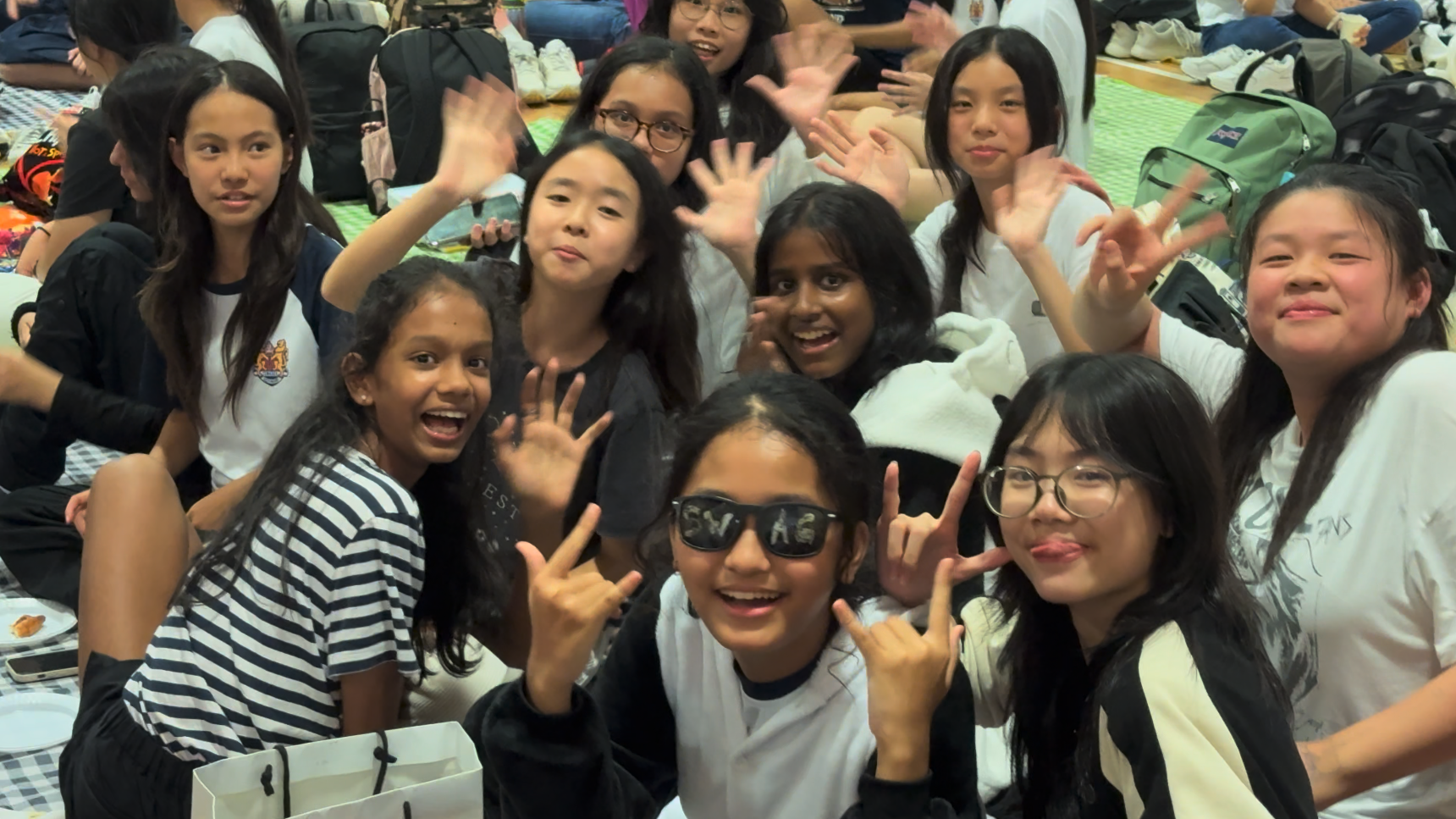 A group of young girls smiling, waving, and making hand gestures at an indoor gathering with backpacks nearby.