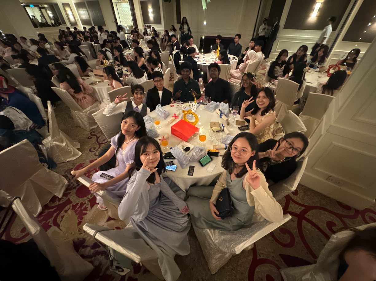 A large group of young people dressed formally sit at round tables in a banquet hall, smiling and posing with peace signs.