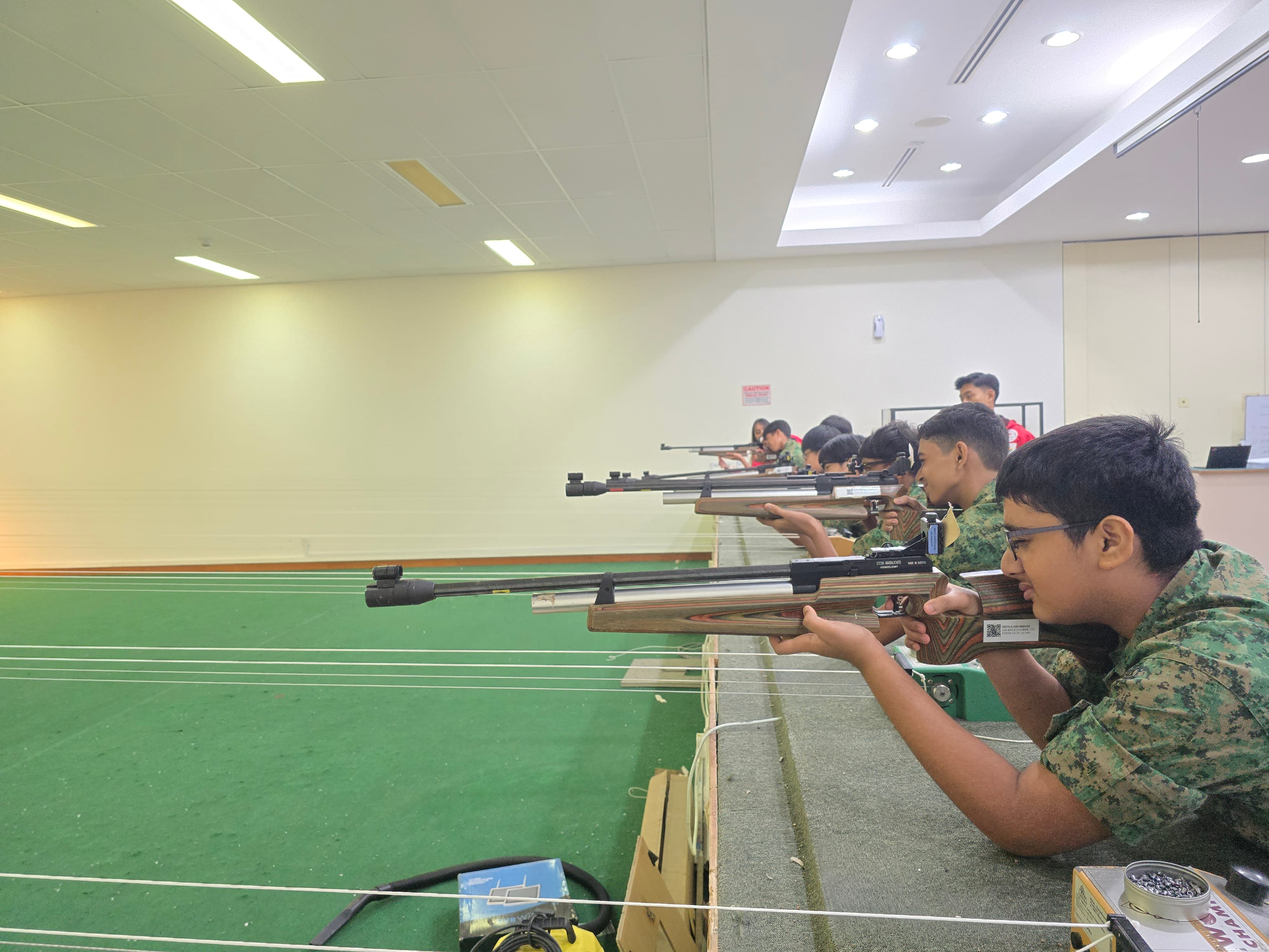 Several young people in camouflage uniforms aim rifles at targets inside an indoor shooting range.