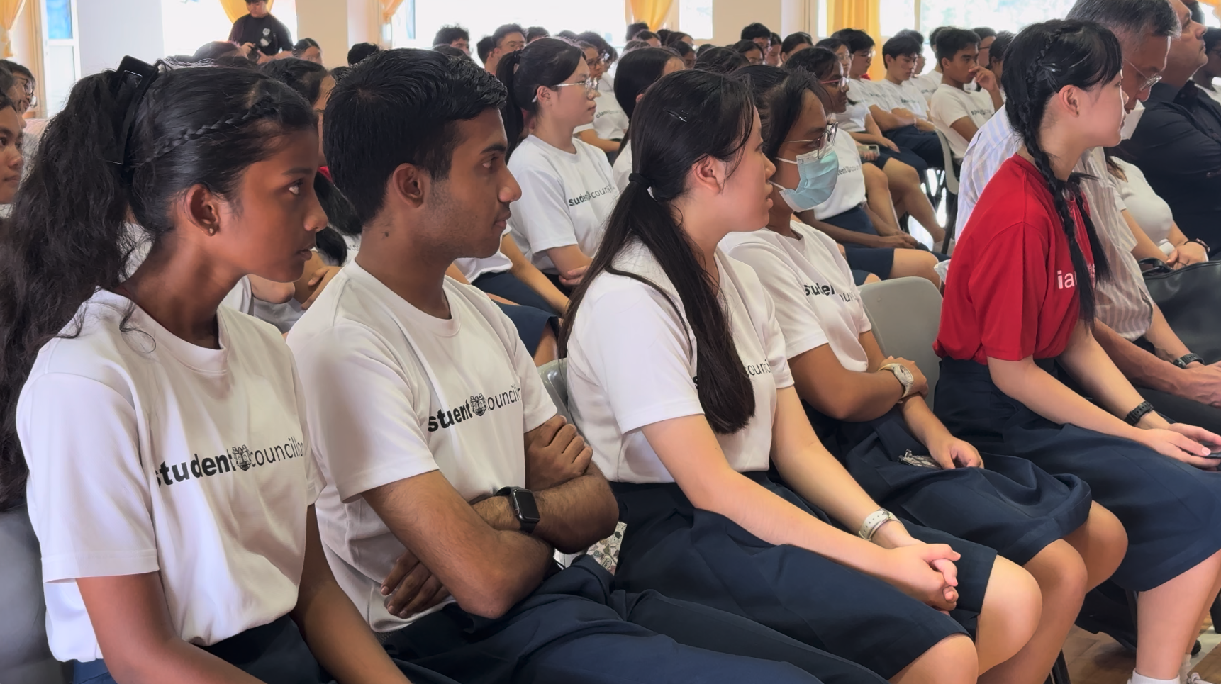 Students in white Student Council shirts sit attentively in rows during a school assembly.