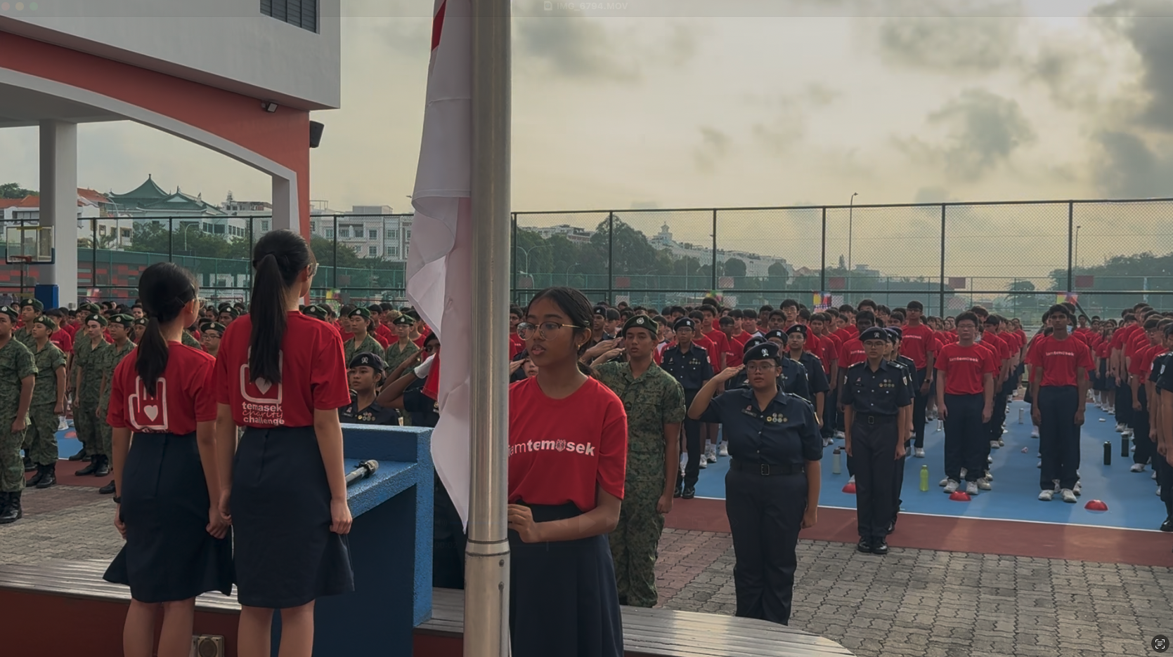 Students in red shirts and uniforms stand at attention during a outdoor flag-raising ceremony at a school courtyard.