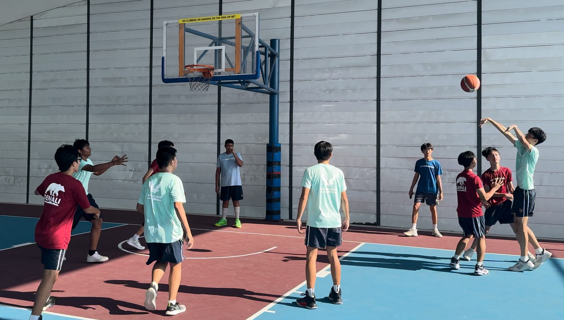 Several young players compete in a basketball game on an outdoor court, with one player shooting toward the hoop.