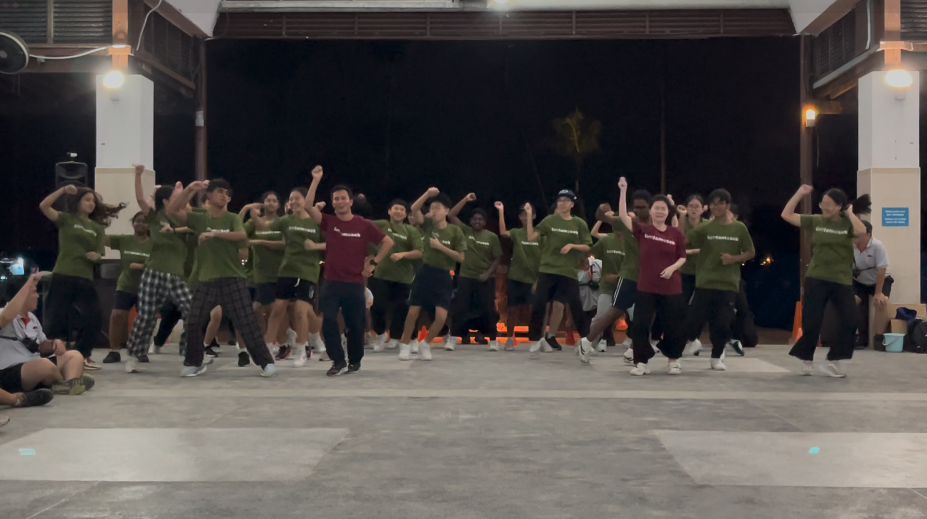 A large group of young people in green and maroon shirts dancing together outdoors at night.