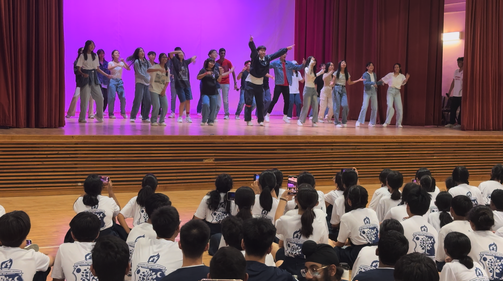 A large group of students dance on a purple-lit stage while seated students in white shirts watch and record with phones.
