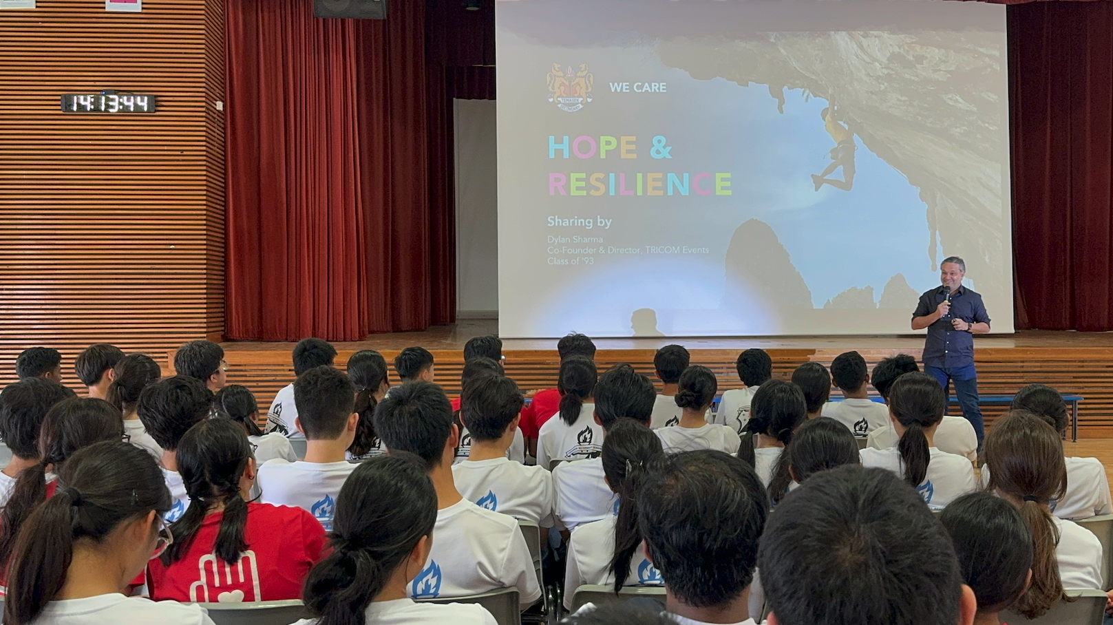 A speaker addresses students in a hall beside a projected slide reading "Hope & Resilience" shared by Dylan Sharma.
