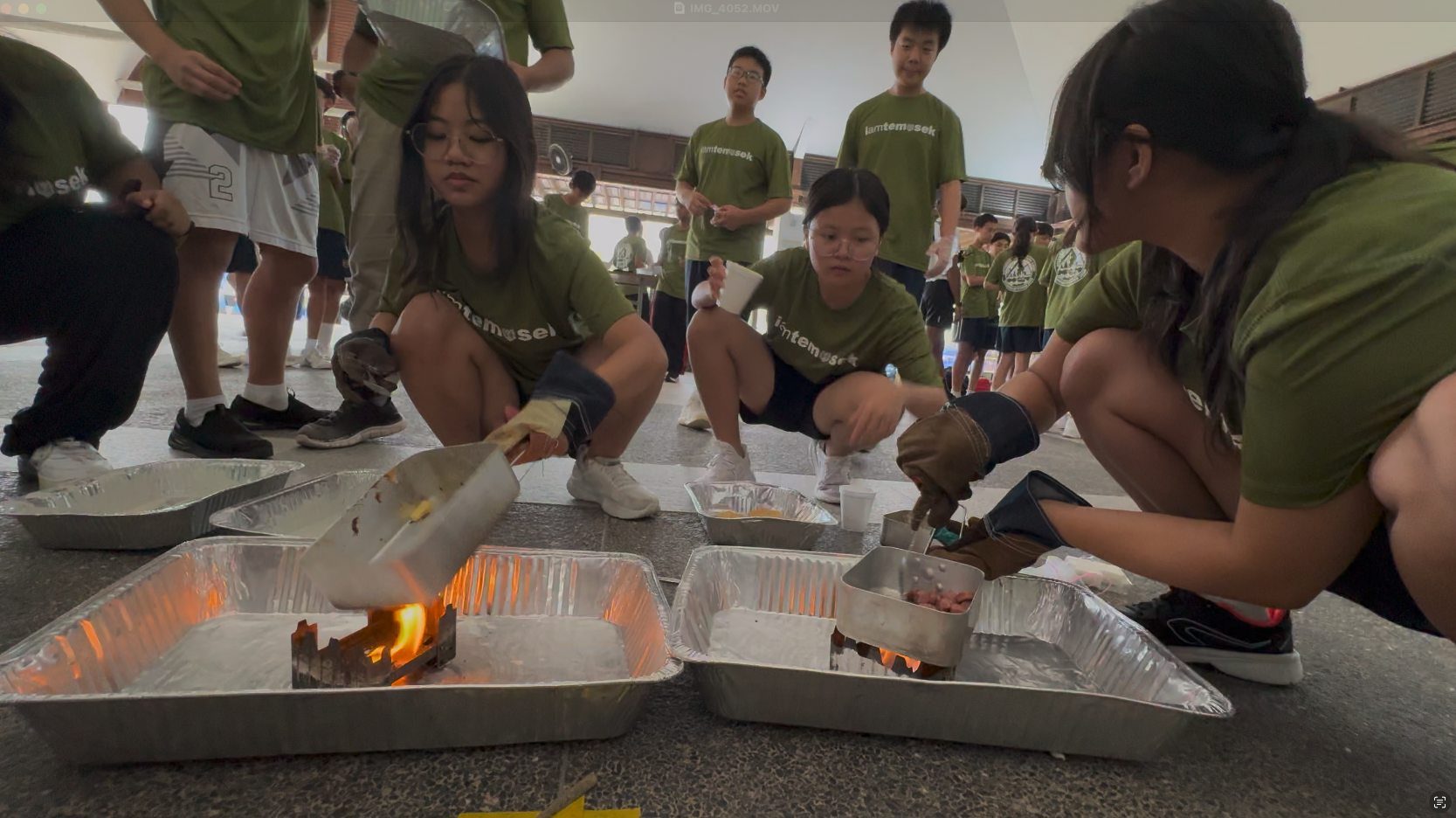 Students in green shirts crouch around foil trays with small fires on a floor during an outdoor activity.