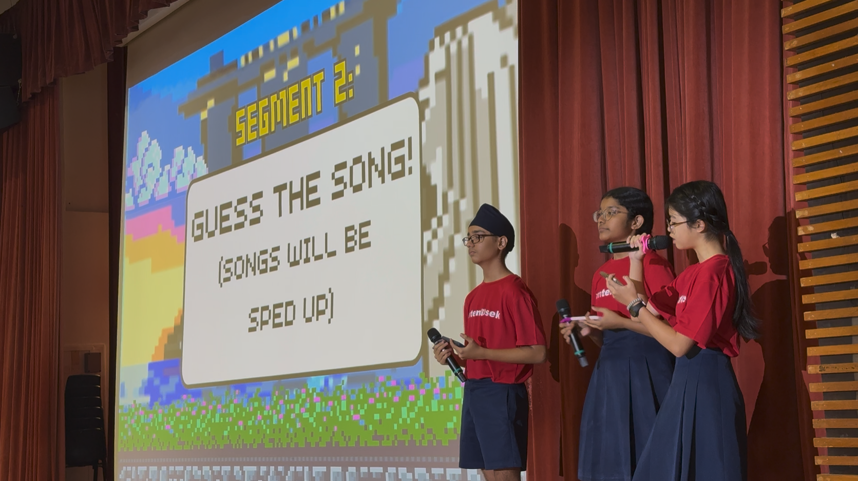 Three students in red shirts hold microphones on stage before a projected slide reading Segment 2: Guess The Song.