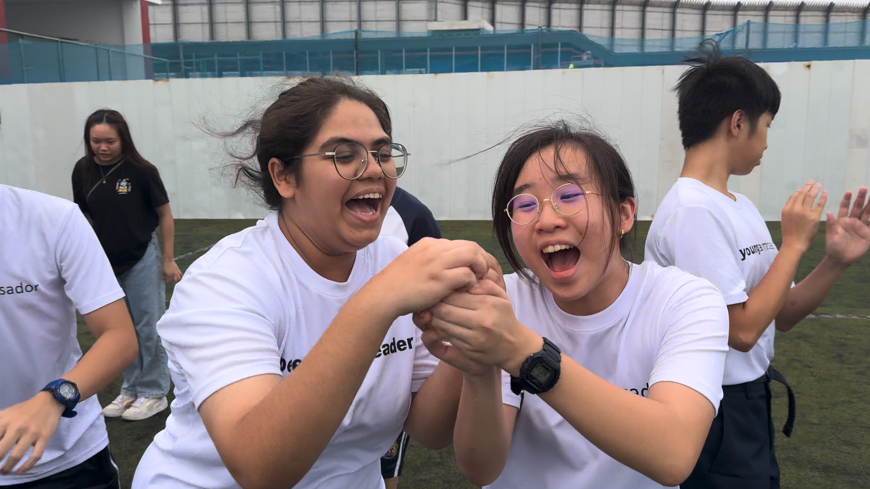 Two young women wearing glasses and white t-shirts laugh and clasp hands together on an outdoor sports field.