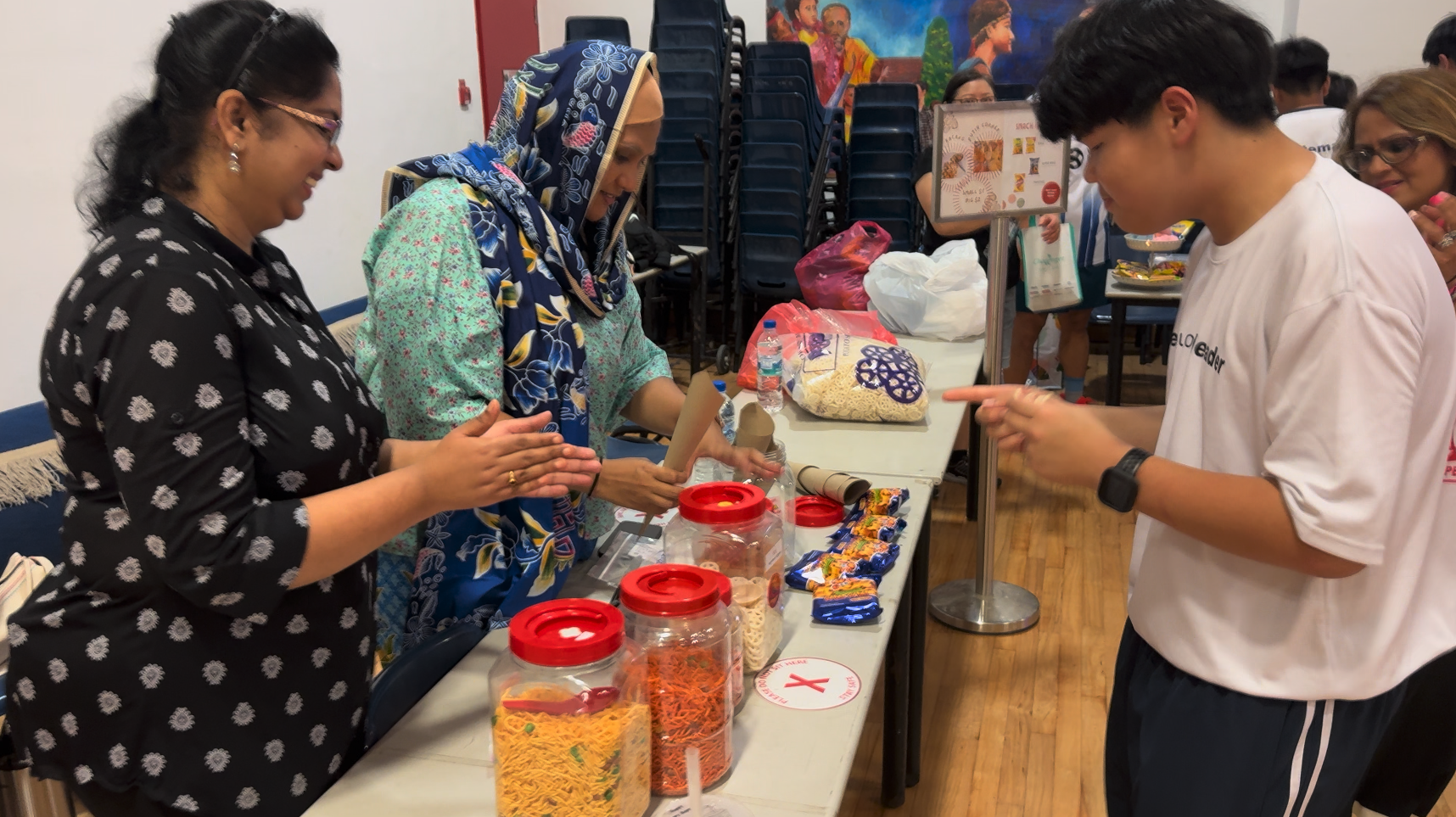 Three people interact at a table with jars of snacks and packaged food in a community hall.