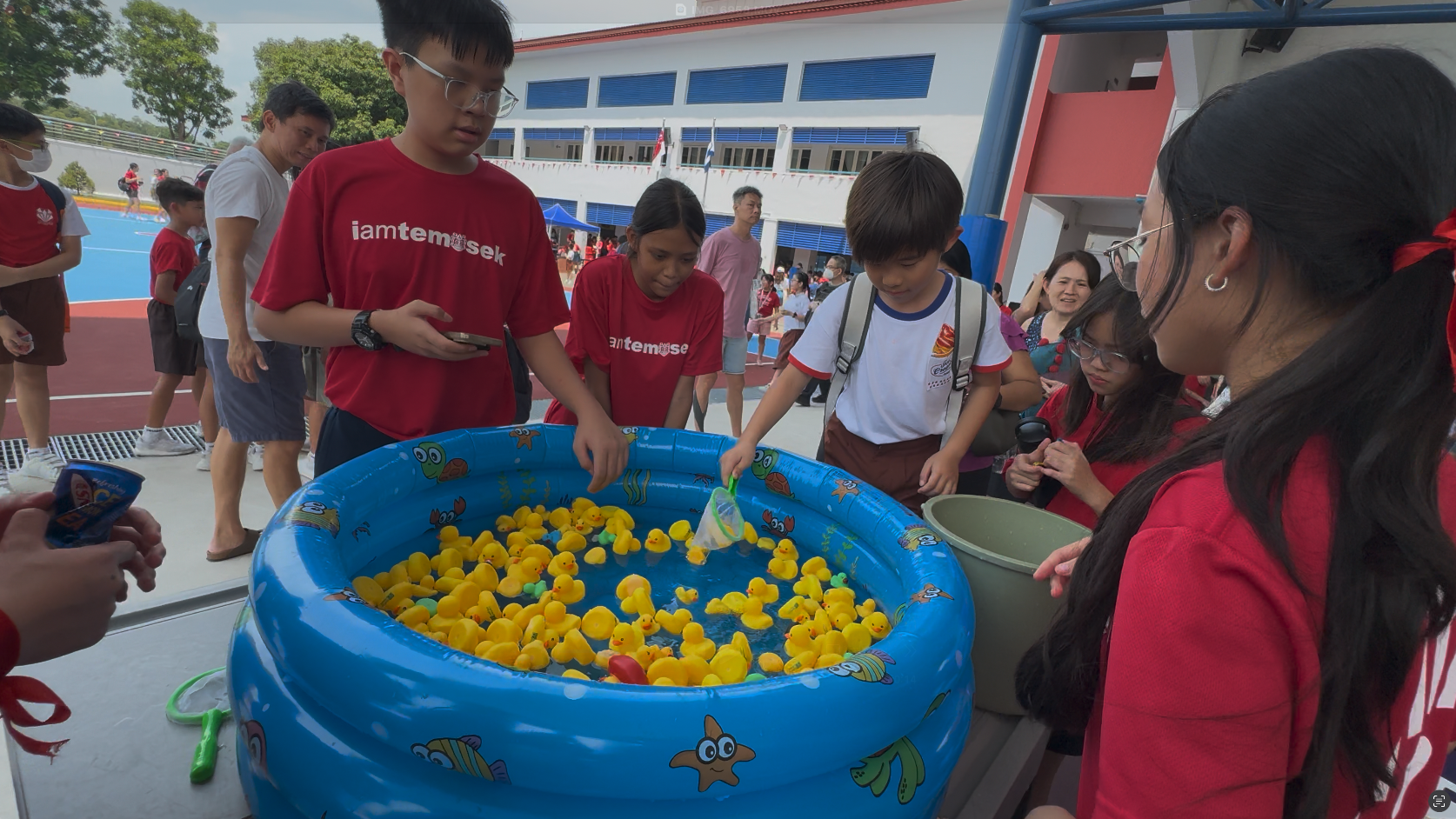 Children in red shirts gather around a blue inflatable pool filled with yellow rubber ducks at an outdoor school event.