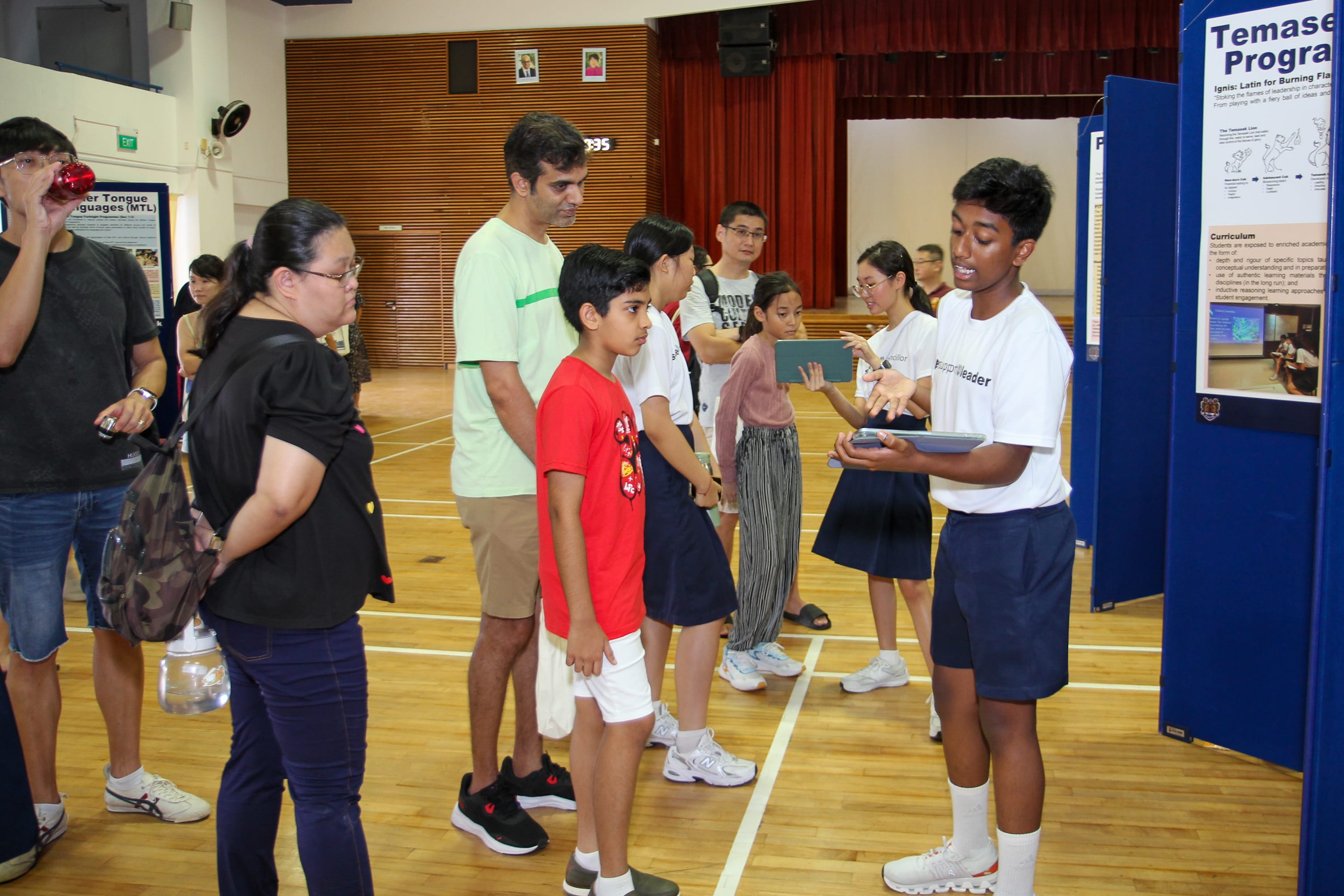 A student in a white shirt presents to a group of adults and children near a school program display board.