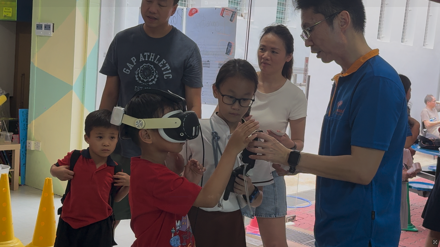 A child wearing a VR headset holds a controller while adults and children gather around indoors.