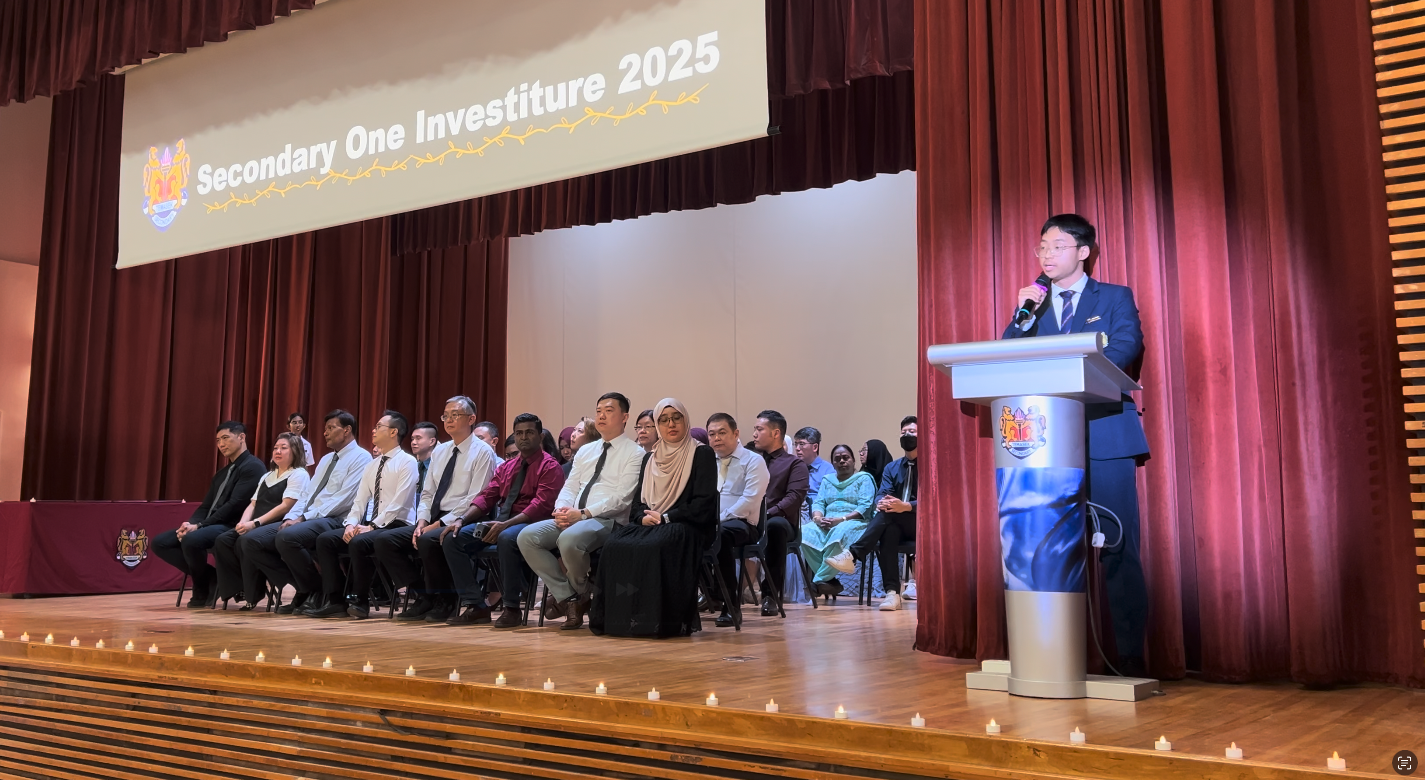 A student speaks at a podium during a Secondary One Investiture 2025 ceremony, with staff seated on stage.